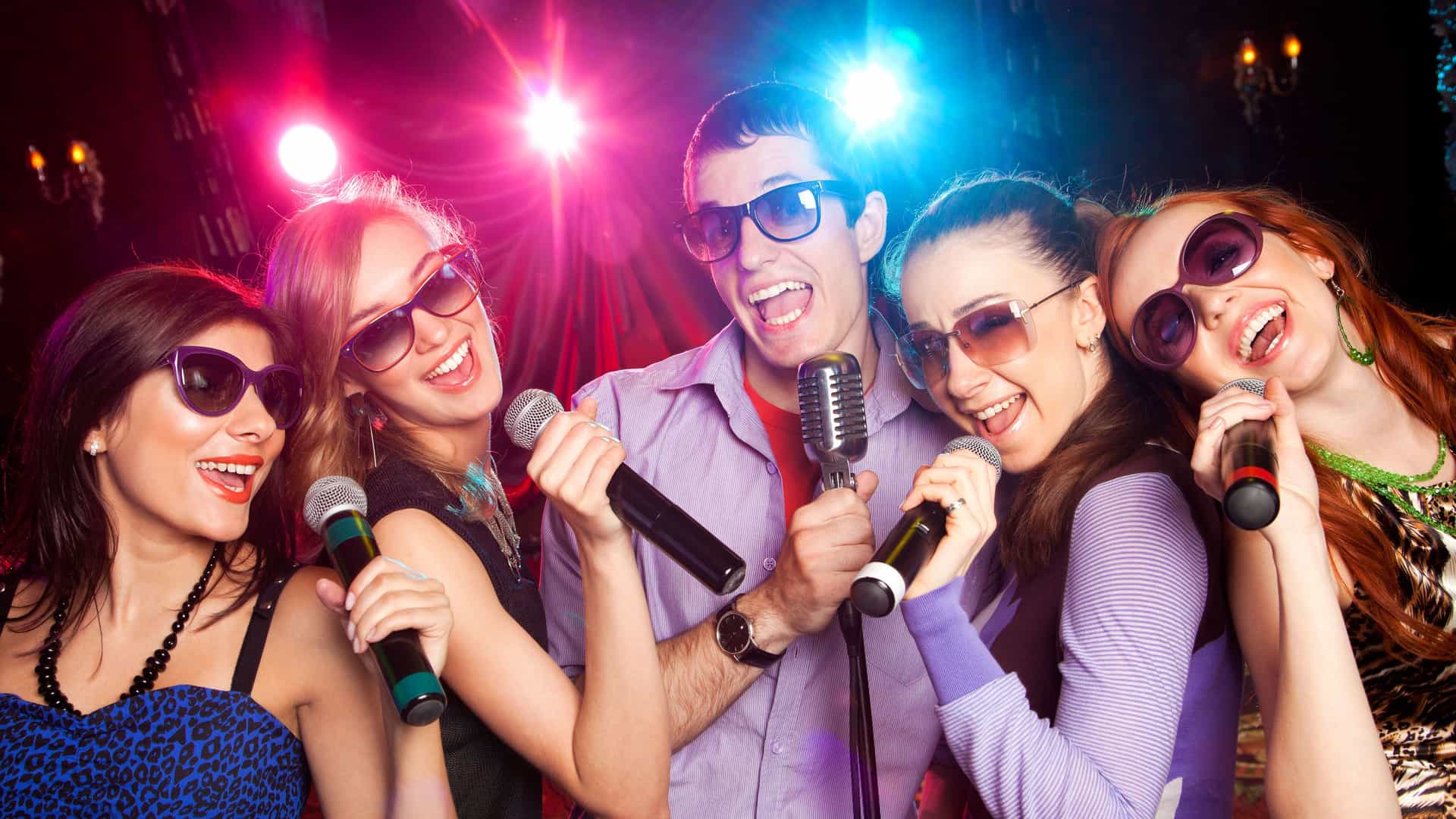 A group of five friends, four women and one man, are singing karaoke together in a dimly lit club. They are holding microphones and smiling brightly. The man in the center is holding a vintage-style microphone on a stand. The group is wearing sunglasses and dressed in stylish casual clothes. The background is dark with bright red and blue spotlights, creating a vibrant and energetic atmosphere. The image represents a popular social activity in the Asian region.