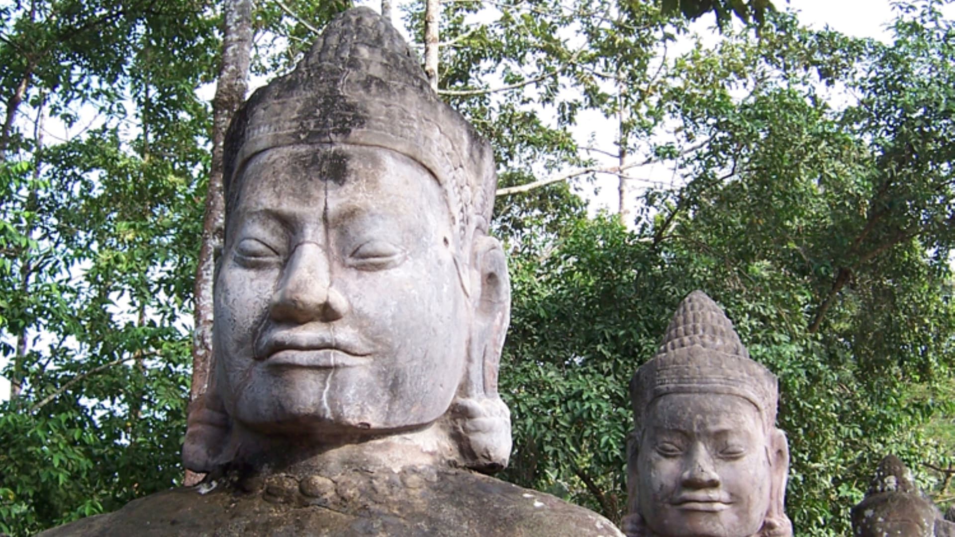 A close-up view of ancient stone carved faces with serene expressions, reminiscent of the Bayon temple statues in Cambodia, set against a backdrop of lush green trees.