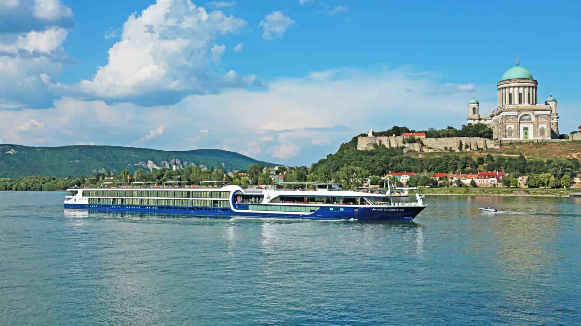 Avalon Waterways river cruise ship sailing on the Danube River past the Esztergom Basilica under a blue sky with white clouds in Esztergom, Hungary.
