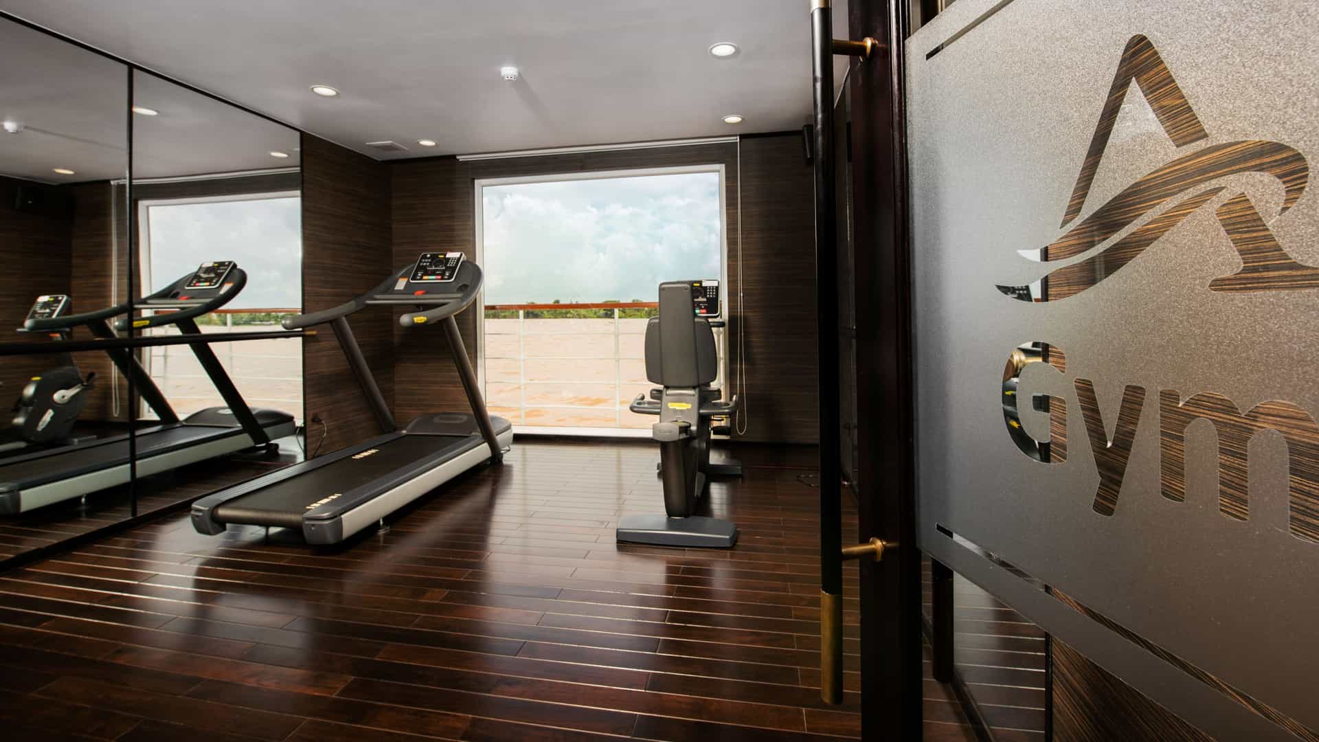 A modern shipboard gym with dark wood flooring, a treadmill, and an exercise bike facing a large window with river views. A glass door in the foreground features a stylized "A" logo and the word "Gym."