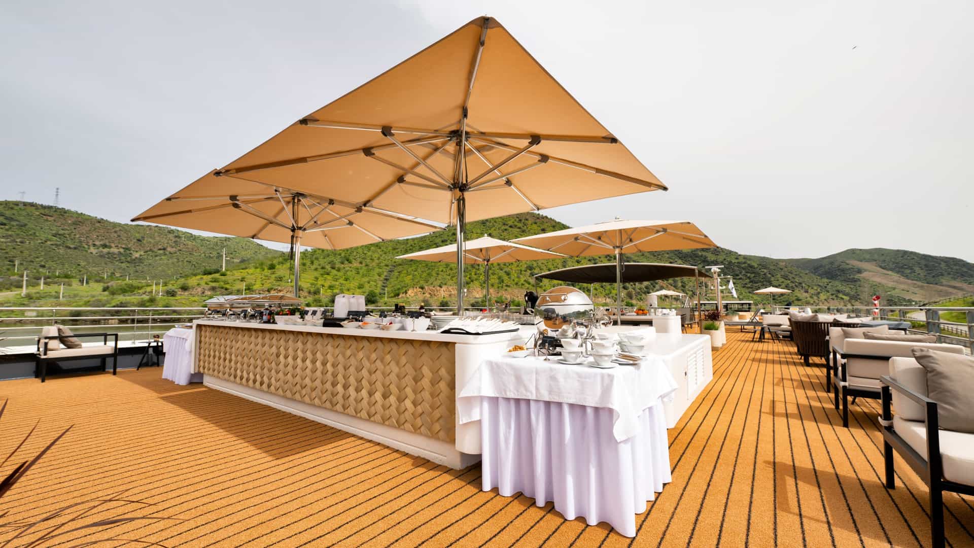 An open-air buffet on a wooden ship deck featuring food stations under large tan umbrellas, lounge seating, and a backdrop of lush green hills.