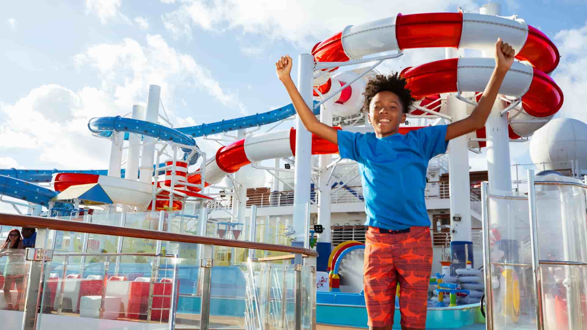 A young boy on the deck of the Carnival Cruise Line's Carnival Miracle ship, standing in front of a colorful water slide, with his hands raised in excitement.