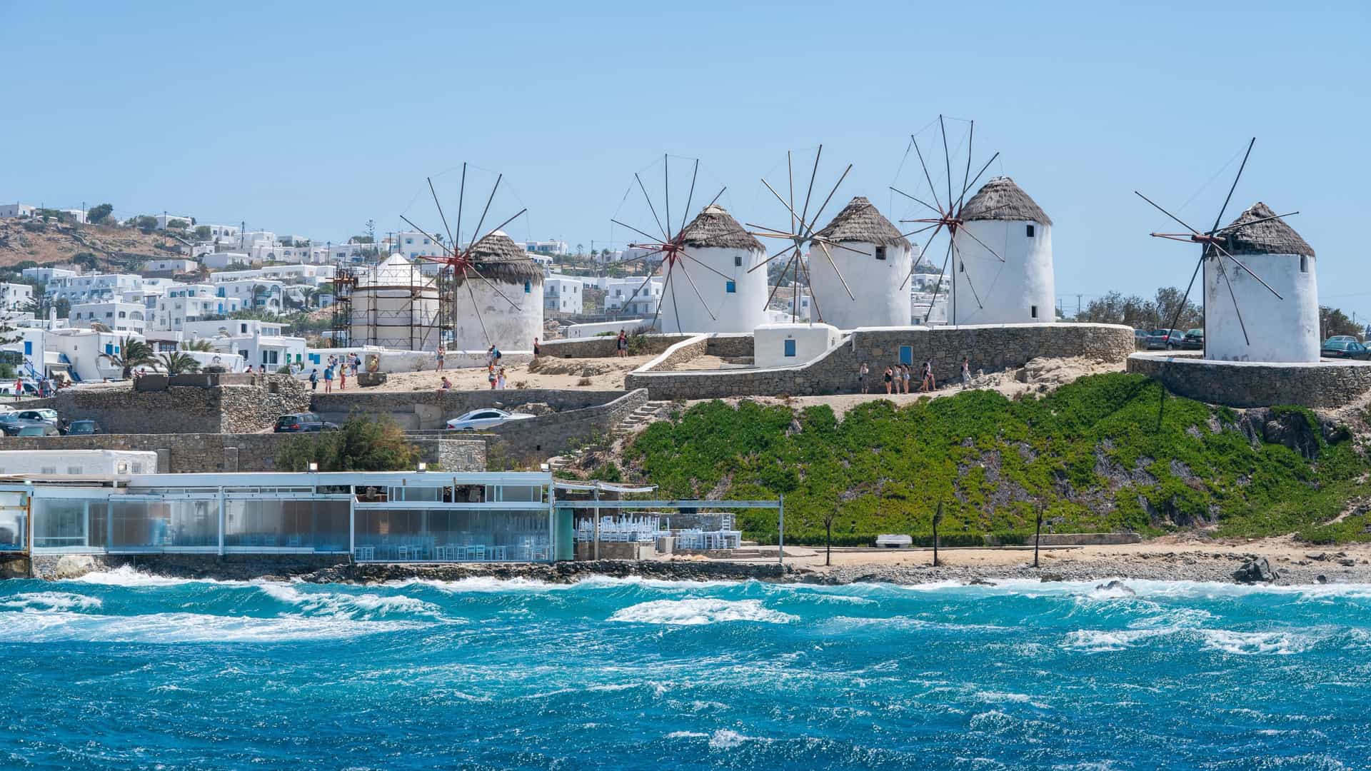 The iconic windmills of Mykonos overlook choppy turquoise waters, with the island's whitewashed village stretching behind them under a clear blue sky.