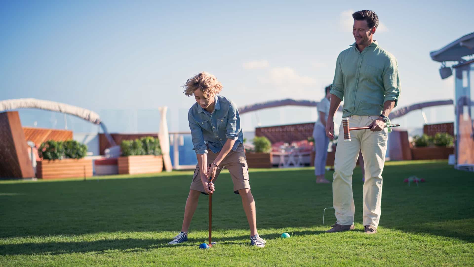 A father and son on the Celebrity Cruises' Celebrity Solstice ship are playing croquet on a manicured lawn, with the boy in a denim shirt and khaki shorts leaning over to hit a ball while his father in a green linen shirt stands nearby and smiles.