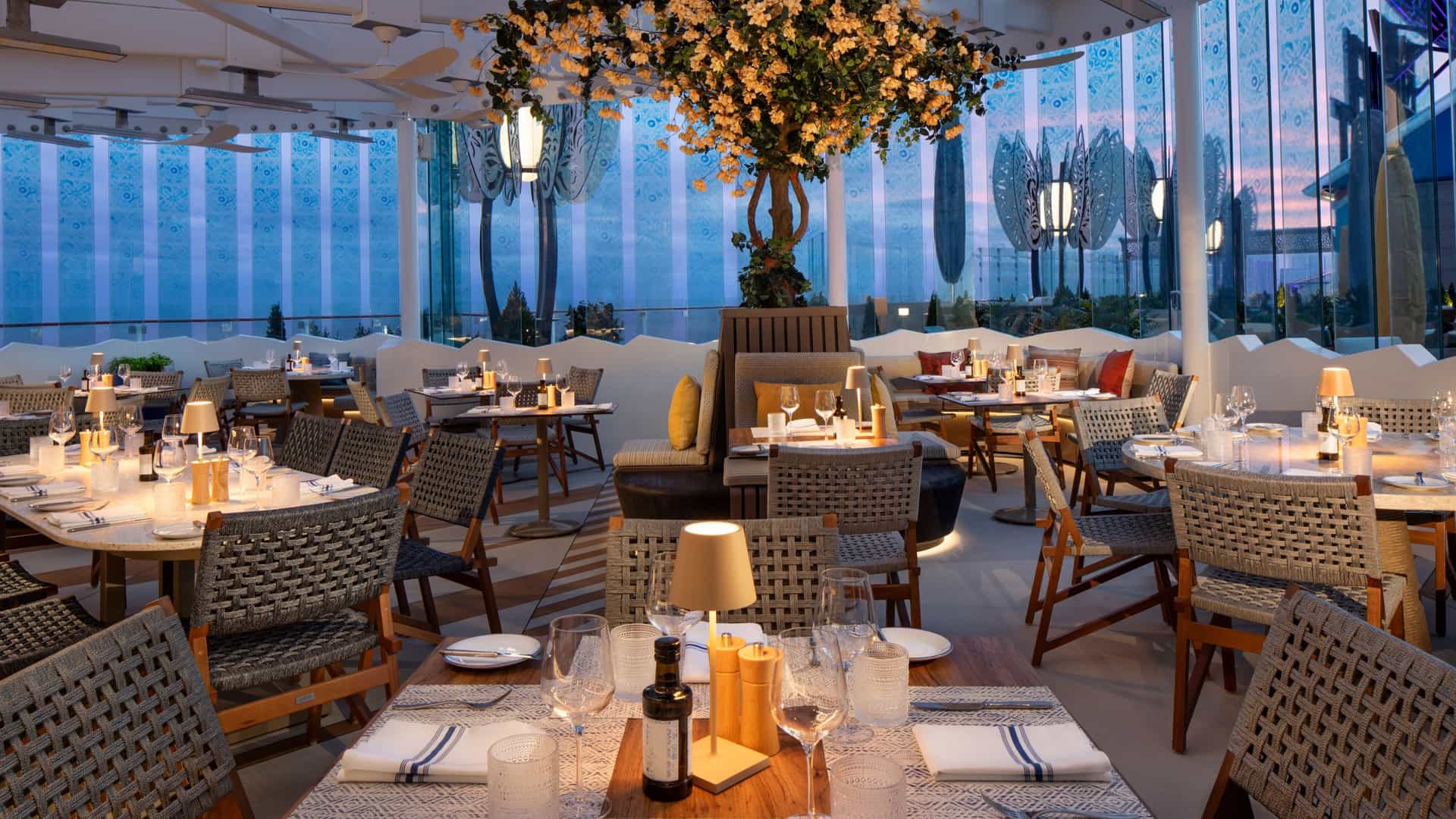 Chic open-air restaurant seating area on Celebrity Xcel featuring stylish tables, warm lighting and a central decorative floral tree.