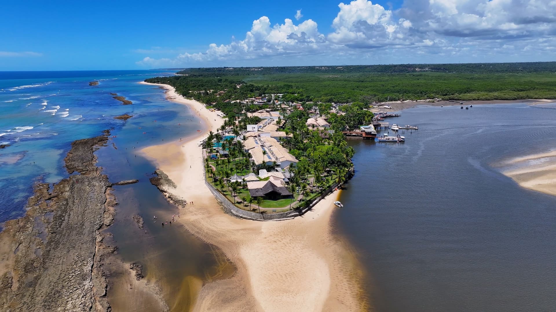 An aerial view of the tropical coastline at Porto Seguro in Bahia, Brazil—a historic South American cruise destination. The image showcases a long, sandy peninsula lined with palm trees and beachside resorts, bordered on one side by a shallow reef in the blue Atlantic Ocean and on the other by a calm river estuary and dense green mangroves under a bright, scattered cloud sky.