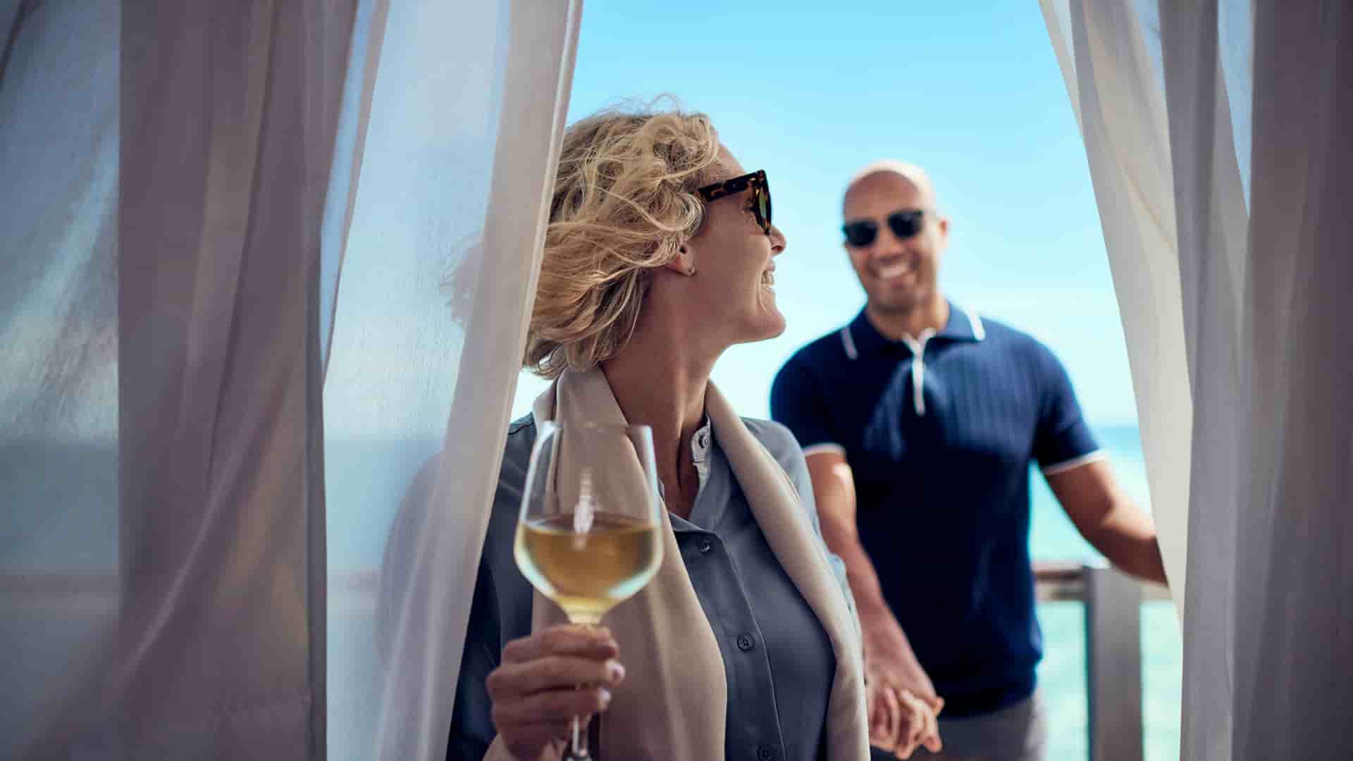 A smiling couple on the Cunard's Queen Victoria ship is standing on their private balcony overlooking the ocean, with the woman holding a glass of white wine while the man holds her hand.