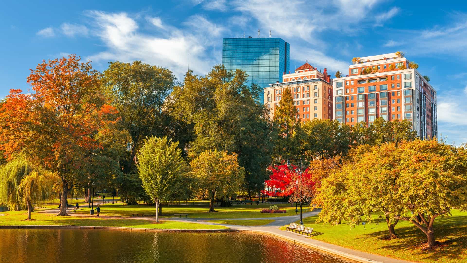 An autumnal view of the Boston Public Garden. The image captures the serene beauty of the park, with a vibrant mix of colorful fall foliage, a calm pond reflecting the trees, and the city skyline visible in the background.