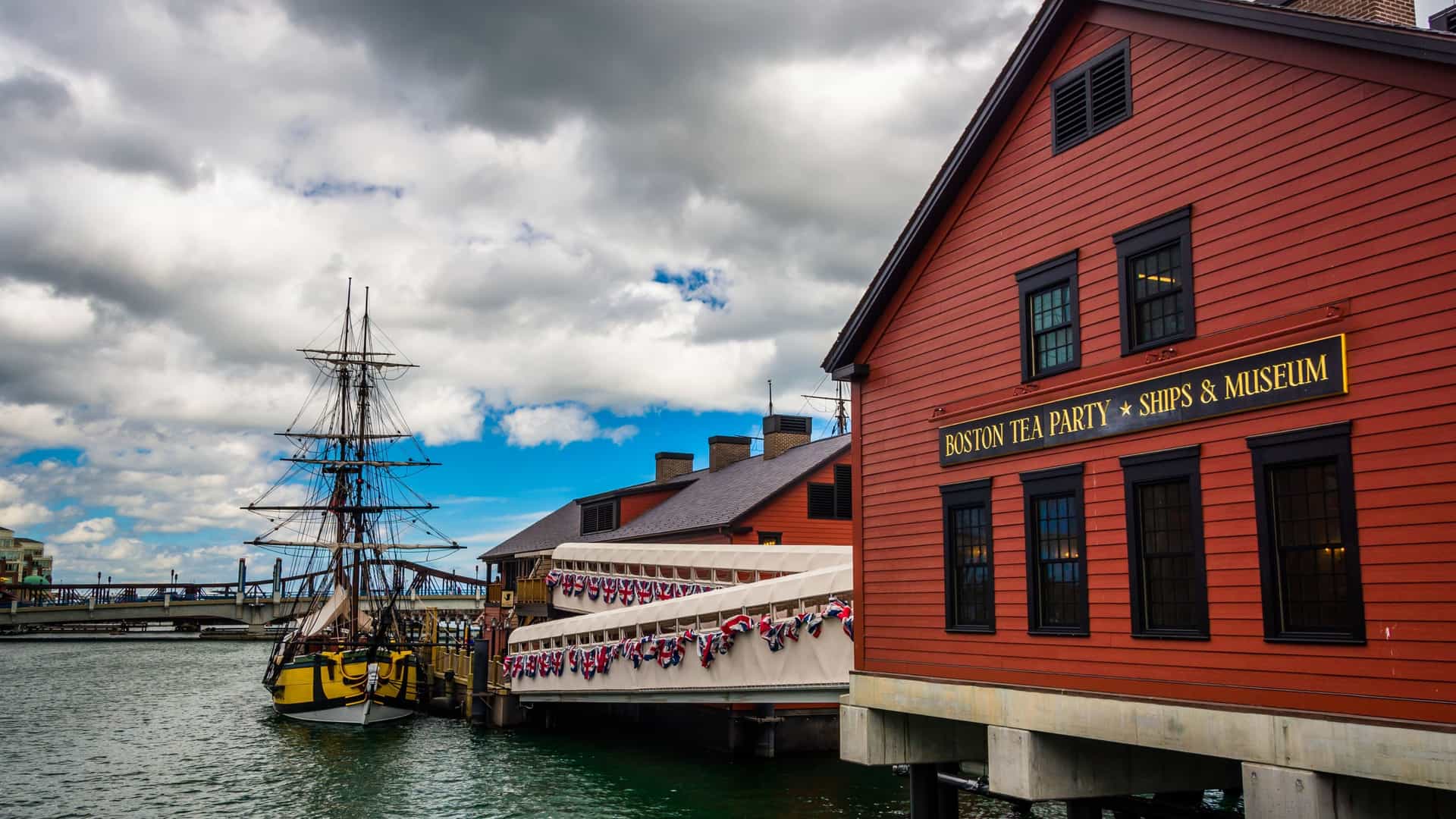 A photograph of the Boston Tea Party Ships & Museum in Boston, Massachusetts. The image shows a replica of an 18th century sailing ship docked on the water, with the Congress Street Bridge and modern city buildings visible in the background.