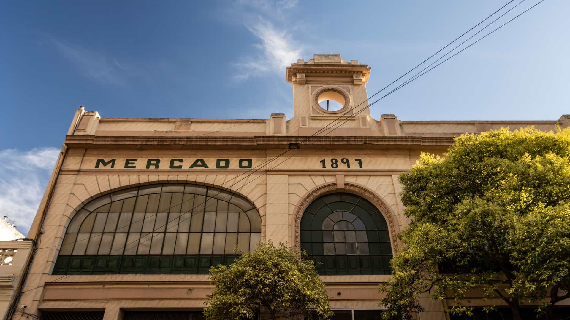 A bright, architectural shot of the interior of the Mercado de San Telmo in Buenos Aires, Argentina. The image showcases the market's historic, industrial age structure, characterized by a vaulted glass and wrought iron ceiling, decorative metal columns, and original iron trusses. Below, the market floor is filled with an eclectic mix of food stalls, modern restaurants, and antique shops, all bathed in natural light filtering through the overhead structure.