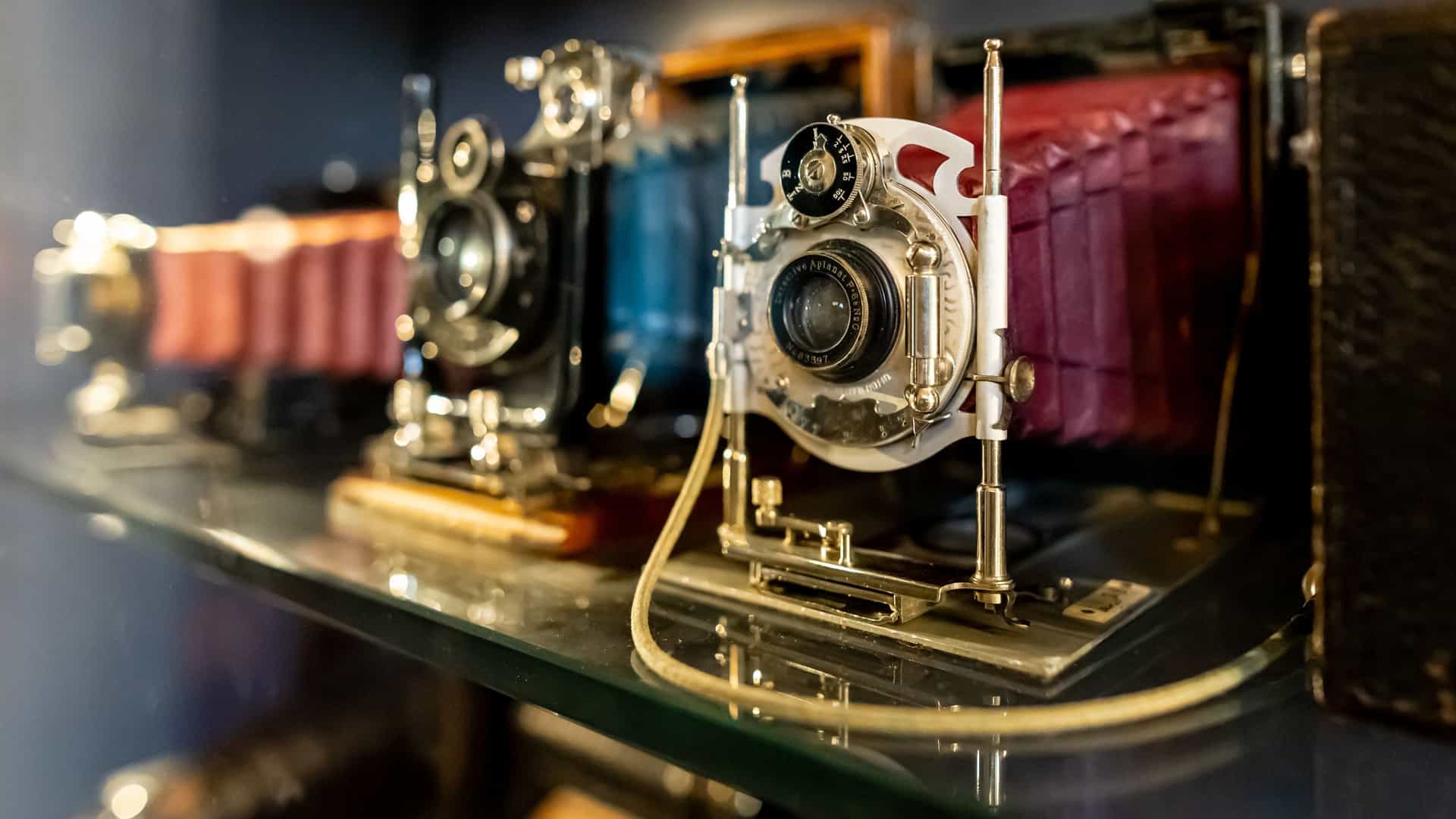 A close up, highly detailed image of two classic, vintage bellows cameras with an antique or leather finish, possibly part of the Musee Nicephore Niepce collection. The focus is on the intricate mechanical components, including the extended black accordion bellows, large lens element, and metal framework, for a still life photograph of antique photographic equipment.