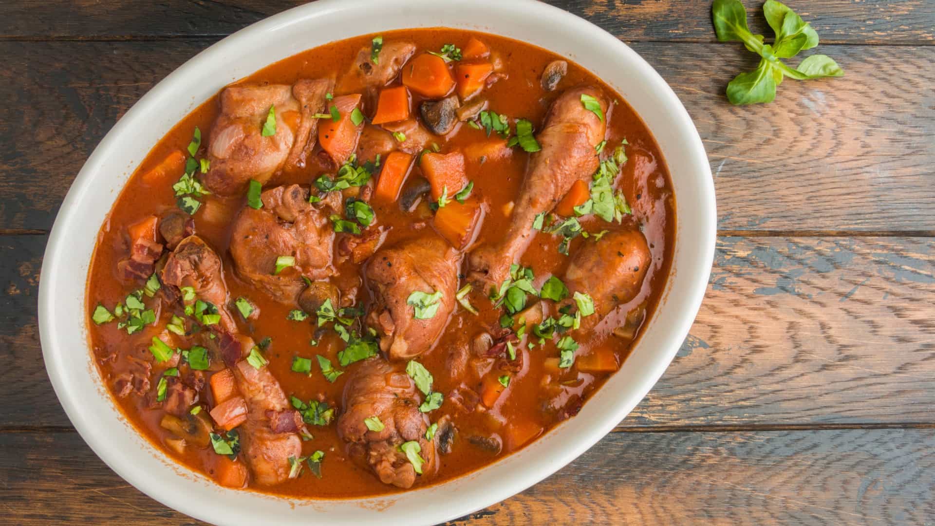 A close up, high angle view of a plate of Coq au Vin, a classic French recipe of chicken braised in a rich, dark red wine sauce with mushrooms and pearl onions. The dish is professionally plated, typically served over mashed potatoes or egg noodles, and garnished with fresh, chopped parsley or herbs.