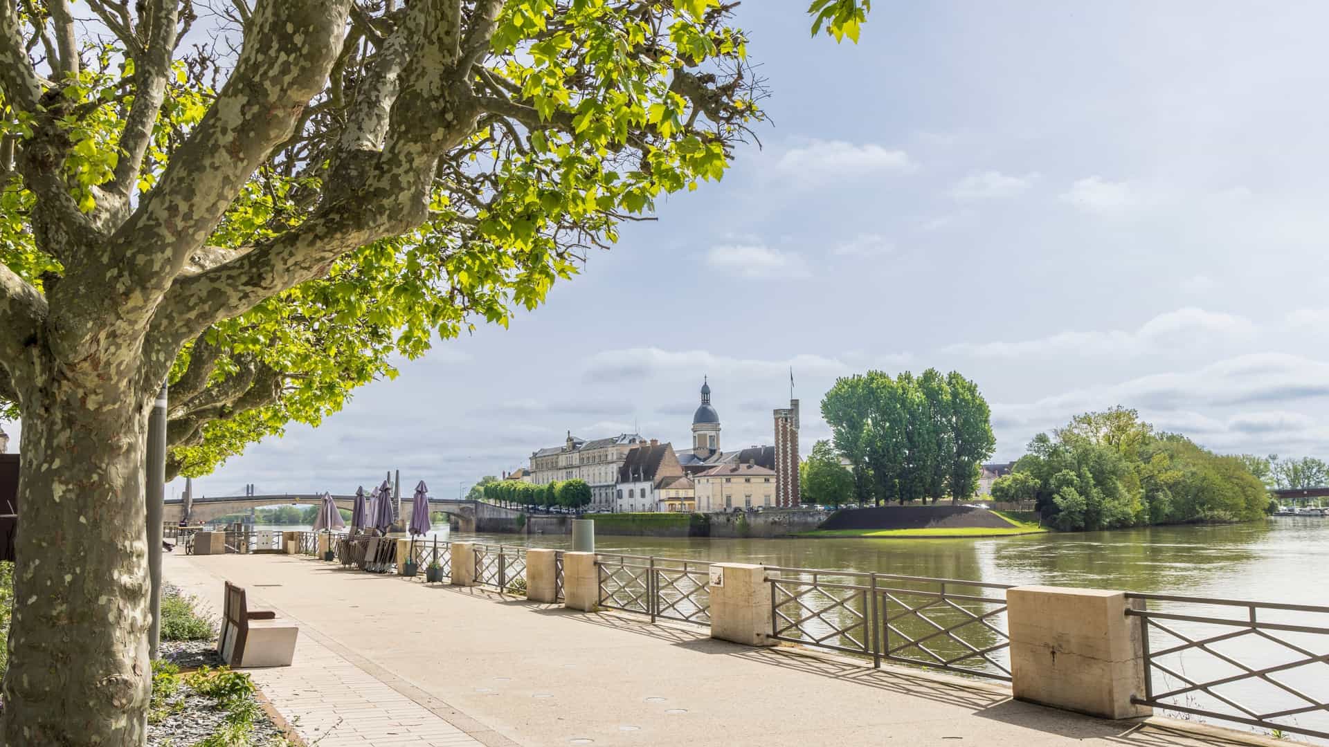 A wide angle view of the Quai des Messageries alongside the Saône River in Lyon, France, featuring a panoramic view of the historic, colorful buildings of the Vieux Lyon district on the opposite bank. The foreground shows the riverbank promenade, highlighting this beautiful area recognized as a UNESCO World Heritage site.