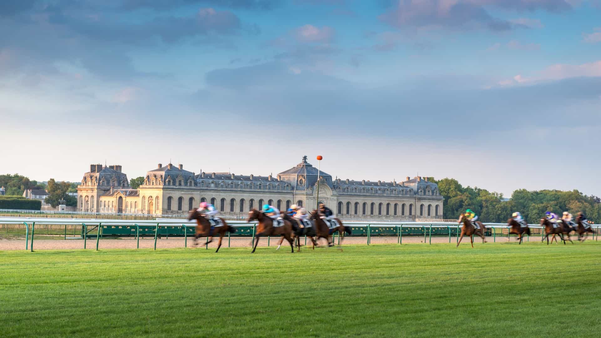 The vast, majestic facade of the Great Stables of Chantilly (Grandes Écuries) is shown, which houses the renowned Living Museum of the Horse (Musée Vivant du Cheval). This stunning 18th century structure is a classical French masterpiece, serving as an architectural landmark and a significant institution dedicated to equestrian culture.