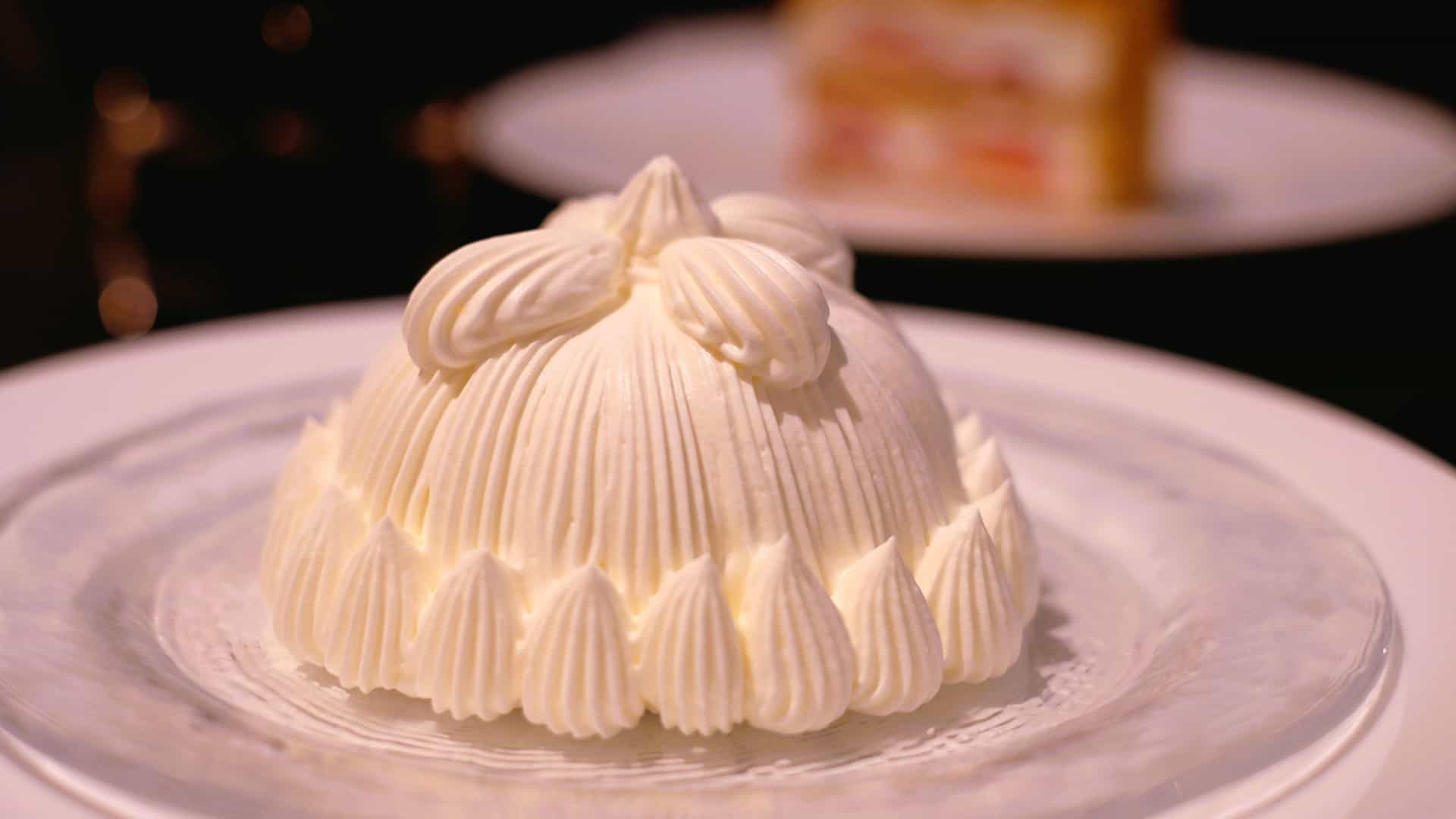 A close up shot of a wedge of elegant layered cake on a white plate, generously topped with smooth, airy crème Chantilly. The slice reveals multiple layers of delicate sponge and cream filling, accented by a light dusting of powdered sugar and a decorative berry.