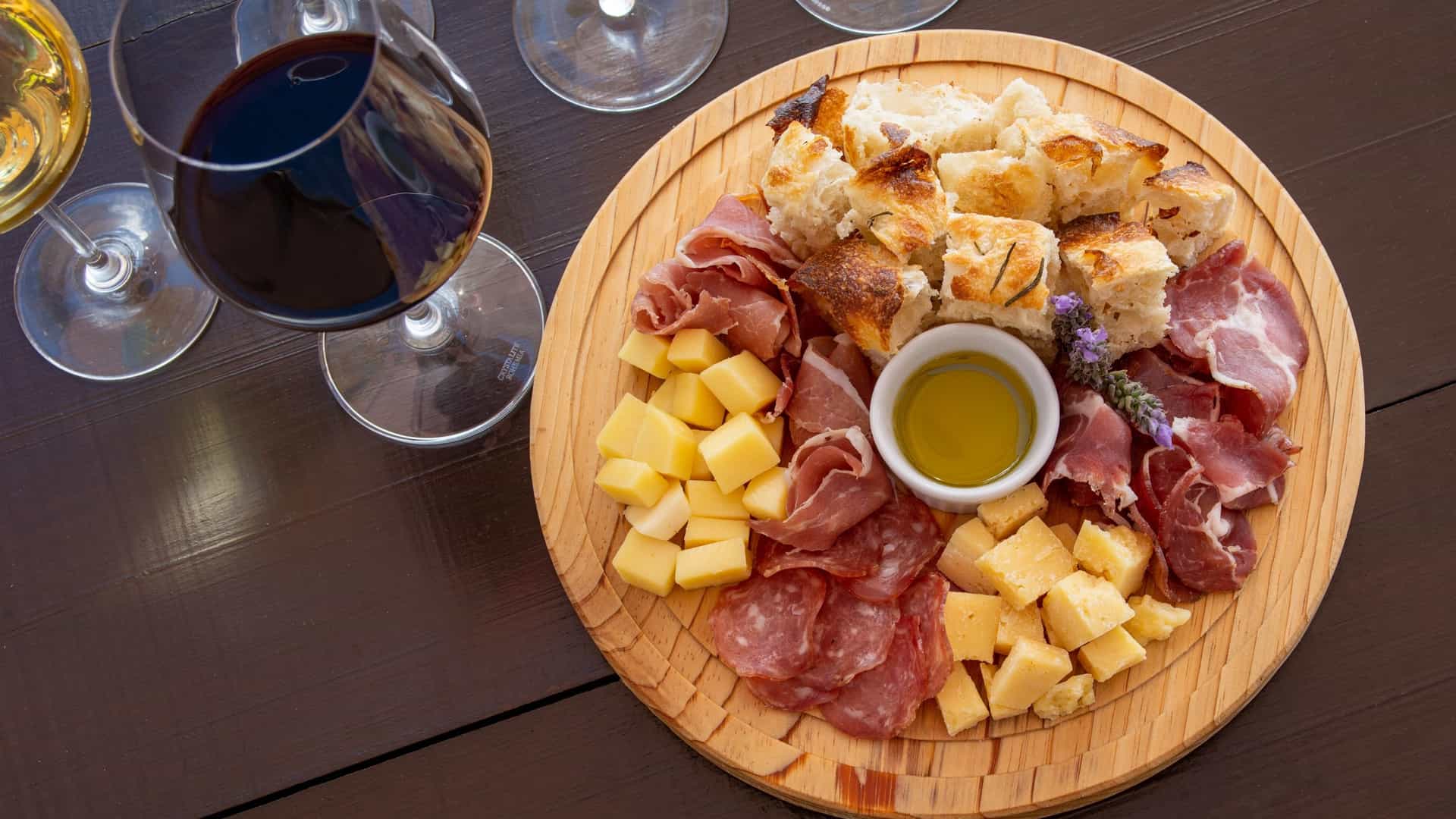 An overhead shot of an appetizing charcuterie board, abundant with sliced cured meats, assorted cheeses, grapes, olives, and breadsticks. The board is elegantly paired with two partially filled glasses of red wine, suggesting a relaxed evening of appetizers and drinks.