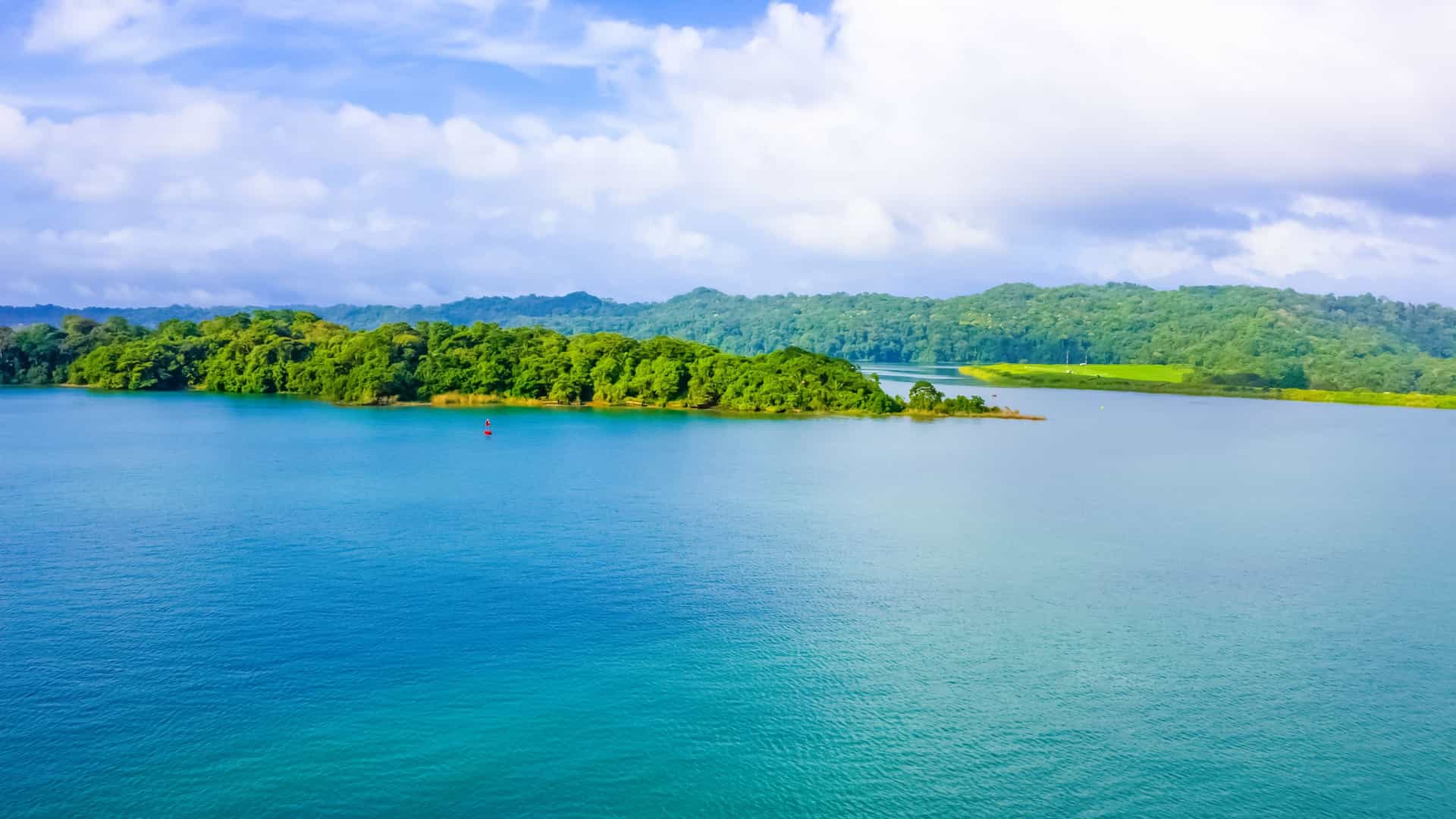 An aerial view of Gatun Lake, a major part of the Panama Canal, with a large expanse of blue green water. The lake is dotted with small, forested islands and surrounded by dense, tropical vegetation under a clear blue sky.