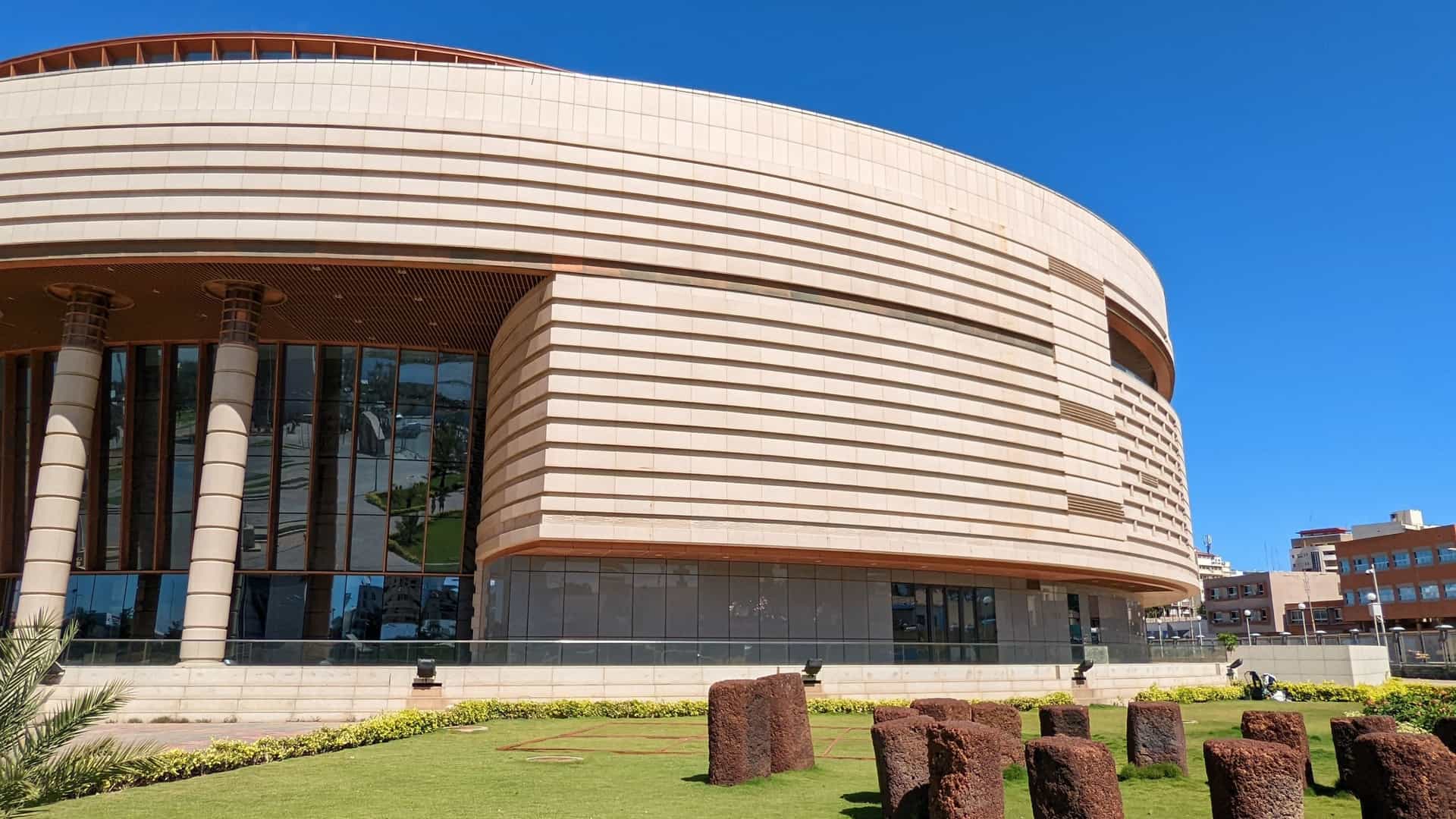 An exterior view of the Museum of Black Civilizations (Musée des civilisations noires) in Dakar, Senegal. The massive building is a modern, circular, ochre colored structure with an upper section inspired by the impluvium architecture of southern Senegal and the ruins of Great Zimbabwe. The image captures the impressive scale of the national museum, which was built with Chinese assistance and is a major cultural symbol in West Africa.