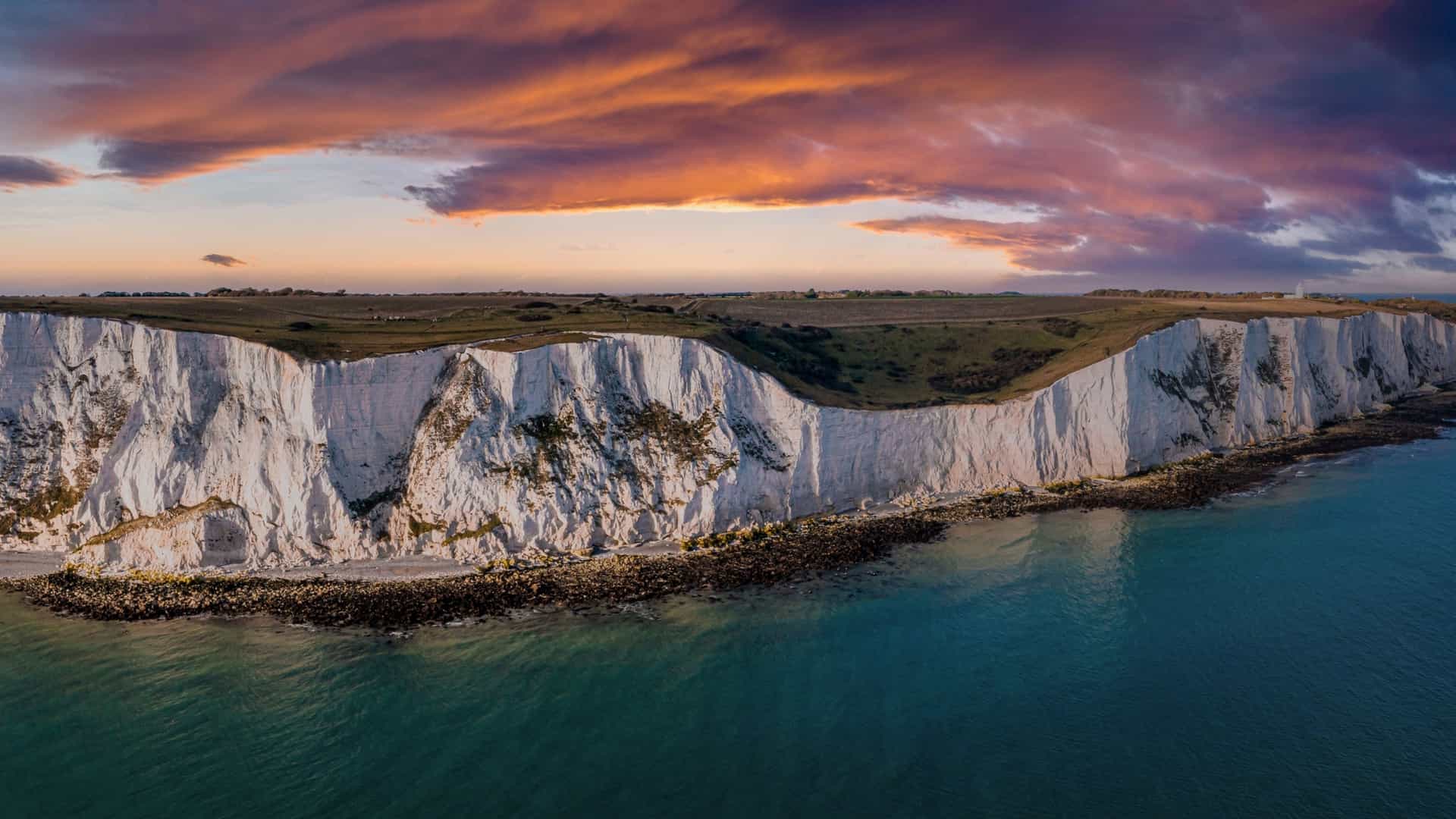 An aerial view of the White Cliffs of Dover, a famous English coastline landmark, overlooking the English Channel. The dramatic chalk cliffs, accented with streaks of black flint, are shown on a sunny day with green fields on top. The image also captures the serene blue water of the English Channel.
