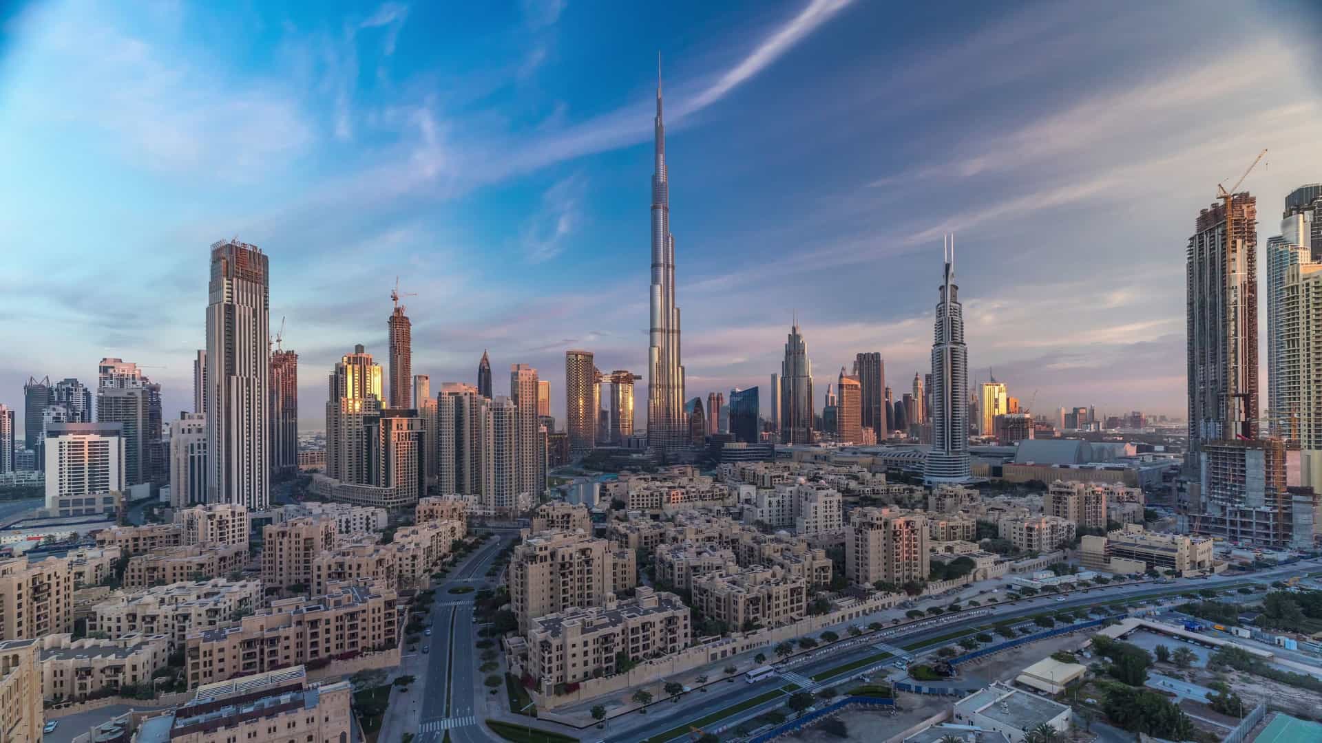 A view of the Dubai downtown skyline at dusk. The image is dominated by the Burj Khalifa, the world's tallest building, which stands prominently amidst a cluster of other modern skyscrapers. The city lights are beginning to illuminate the buildings and streets below.