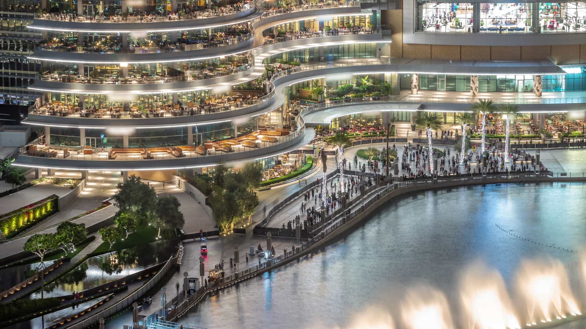 A high angle, nighttime photograph of the tiered dining and retail complex on the promenade outside the Dubai Mall. The modern building features multiple illuminated levels of outdoor seating, overlooking the Dubai Fountain Lake where a crowd is gathered along the waterfront, with bright fountain jets visible in the lower right.