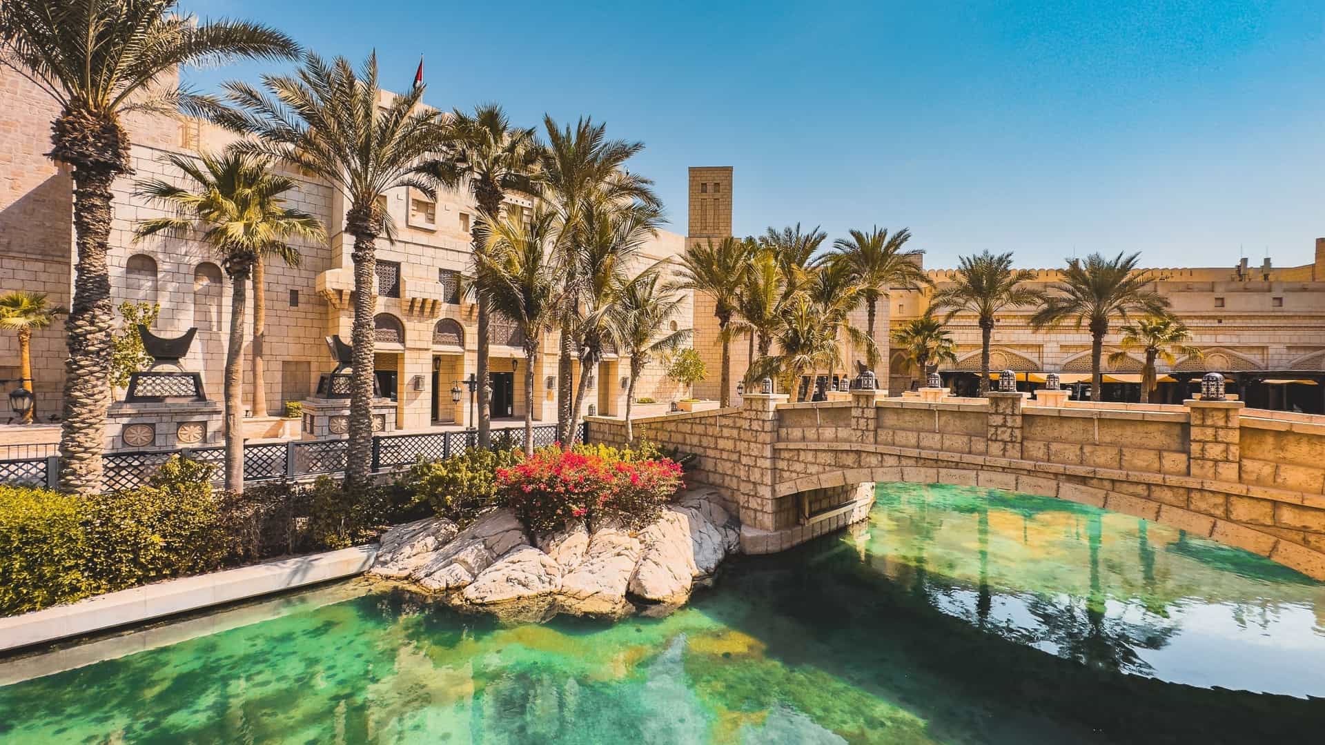 A traditional Arabian style marketplace, Souk Madinat Jumeirah, in Dubai. The image shows a serene water canal with a traditional abra boat, surrounded by buildings with ornate archways and wind towers. The iconic Burj Al Arab hotel is visible in the background.