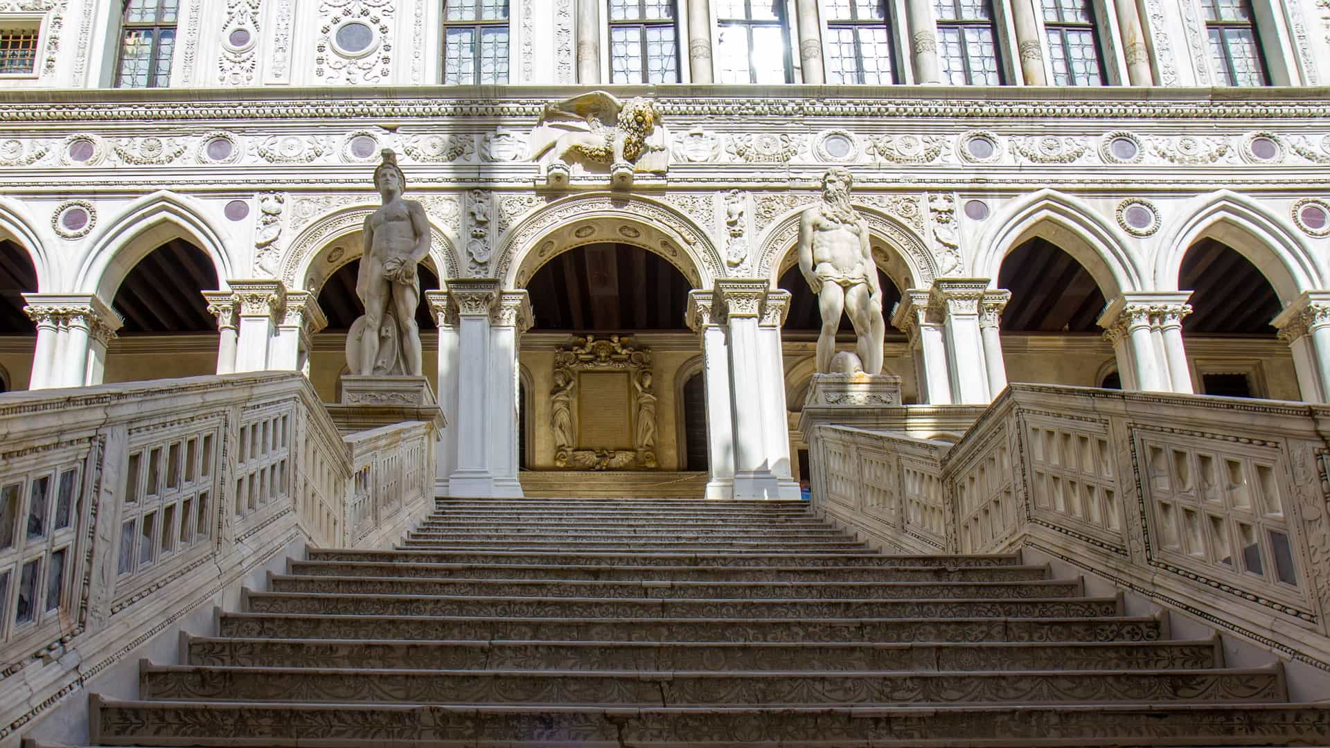 A view of the inner courtyard of the Doge's Palace in Venice, Italy. The courtyard is surrounded by the palace's two main wings, with the Gothic style architecture on one side and Renaissance architecture on the other. The image highlights the detailed marble facades, ornate archways, and the famous Giants' Staircase leading up to the palace.