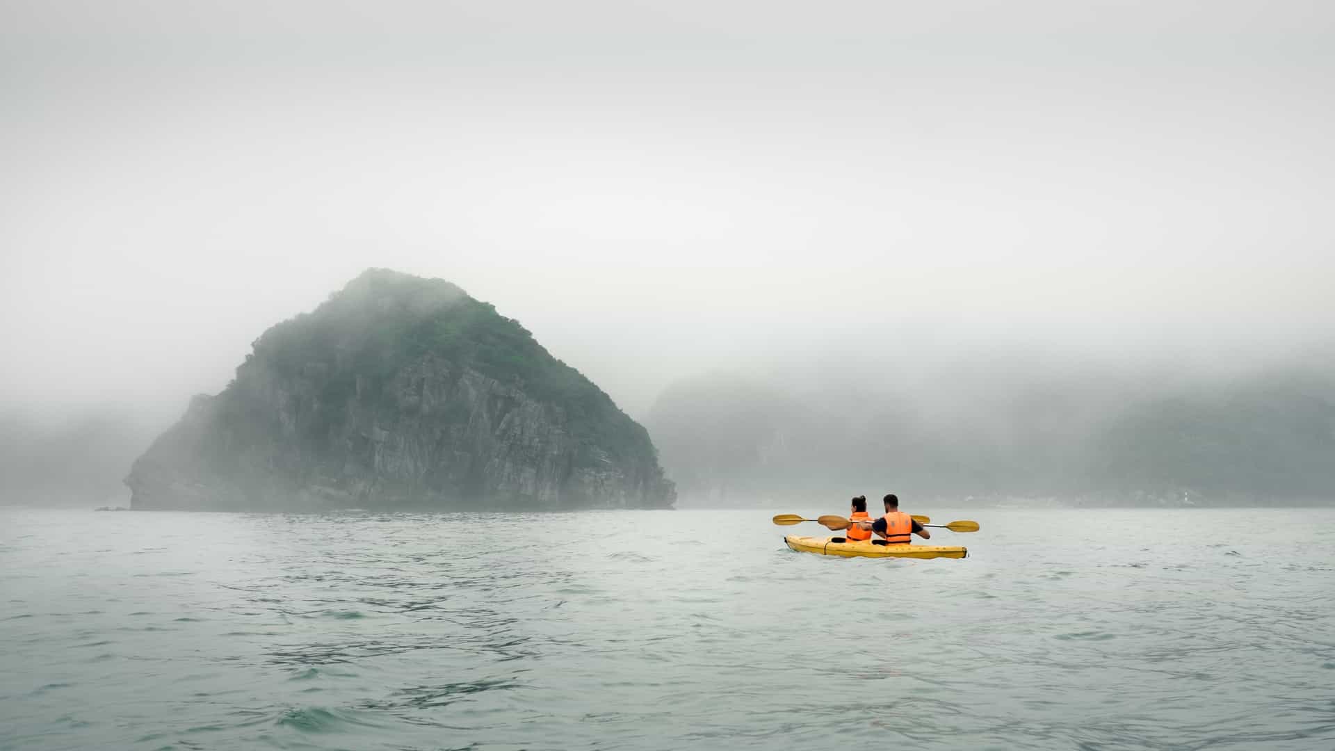 A couple paddles a red kayak through a calm, foggy sea in Ha Long Bay, Vietnam. The water reflects the hazy, overcast sky. Towering, mist shrouded limestone karsts rise dramatically in the background, their silhouettes creating a serene and mysterious atmosphere. The couple is shown from behind, both wearing life vests, with the person in the back holding a paddle.