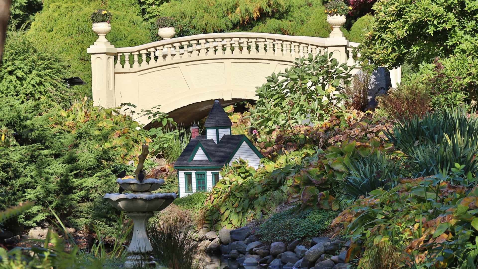 A view of the Francis Fitzgerald Bridge in the Halifax Public Gardens. The concrete bridge, with ornate balusters and decorative urns, spans a calm pond. The surrounding area is lush with green trees and manicured lawns, typical of a Victorian era garden.