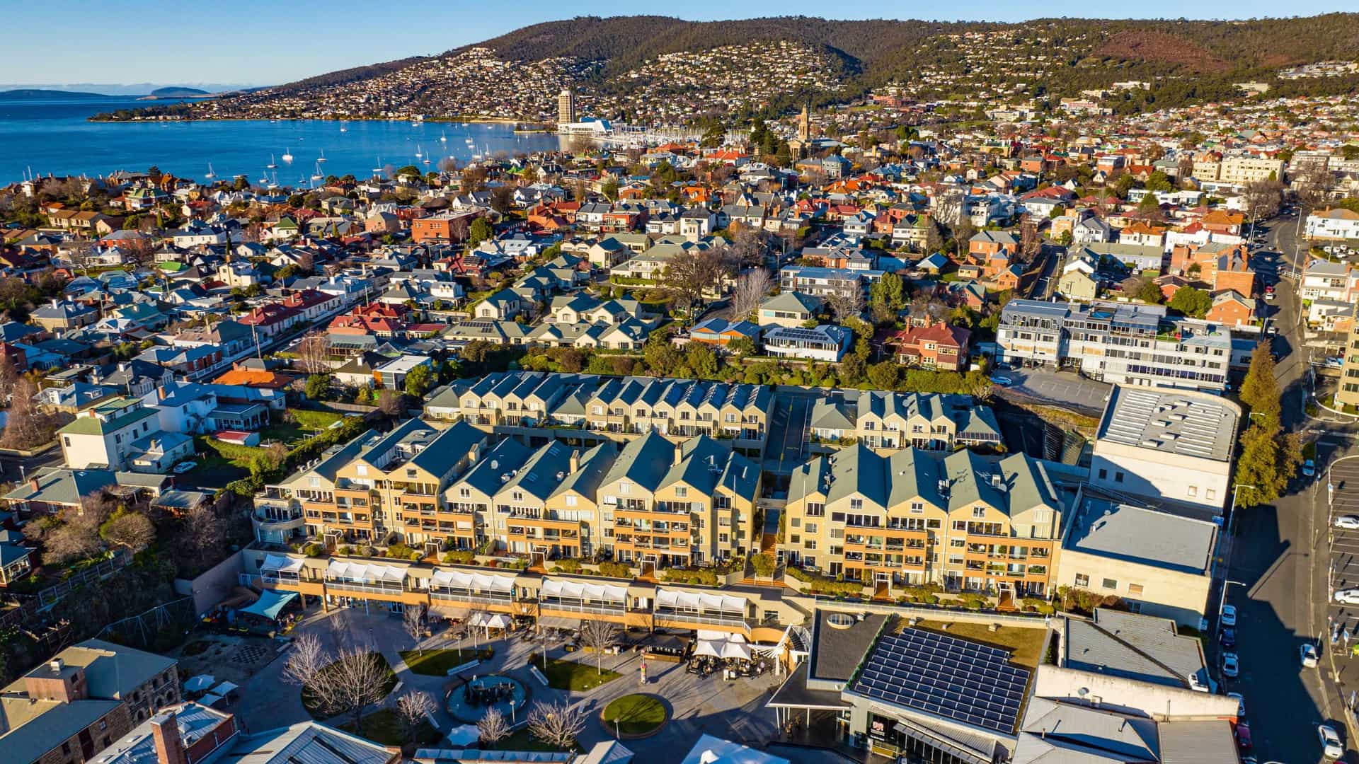 An aerial view of Hobart, Tasmania, featuring the historic areas of Salamanca Place and Battery Point. The image captures the waterfront's distinctive long row of Georgian sandstone warehouses at Salamanca Place and the tightly-packed, colonial-era cottages and streets of Battery Point, with Mount Wellington visible in the distance.
