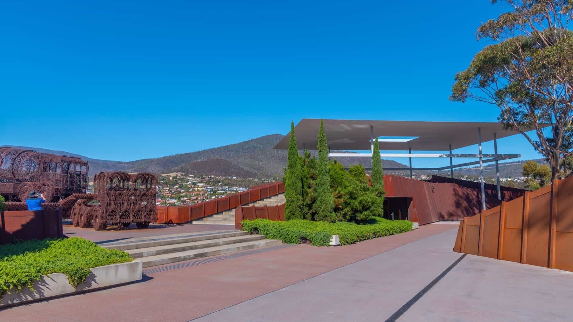 An exterior view of the MONA (Museum of Old and New Art), located on the banks of the Derwent River near Hobart, Tasmania, Australia. The image captures the modern, low-profile architecture of the museum, which is mostly subterranean, situated on the Moorilla Peninsula, with surrounding vineyards and the river visible in the background.