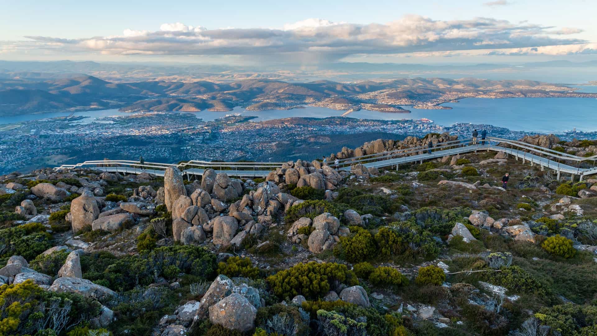 A photograph of the summit of Mount Wellington, also known as Kunanyi, near Hobart, Tasmania. The image is taken during a sunset, casting an orange and pink glow across the sky. The rocky, rugged terrain of the mountain's summit is visible in the foreground, with the city of Hobart and the surrounding landscape stretching out below.