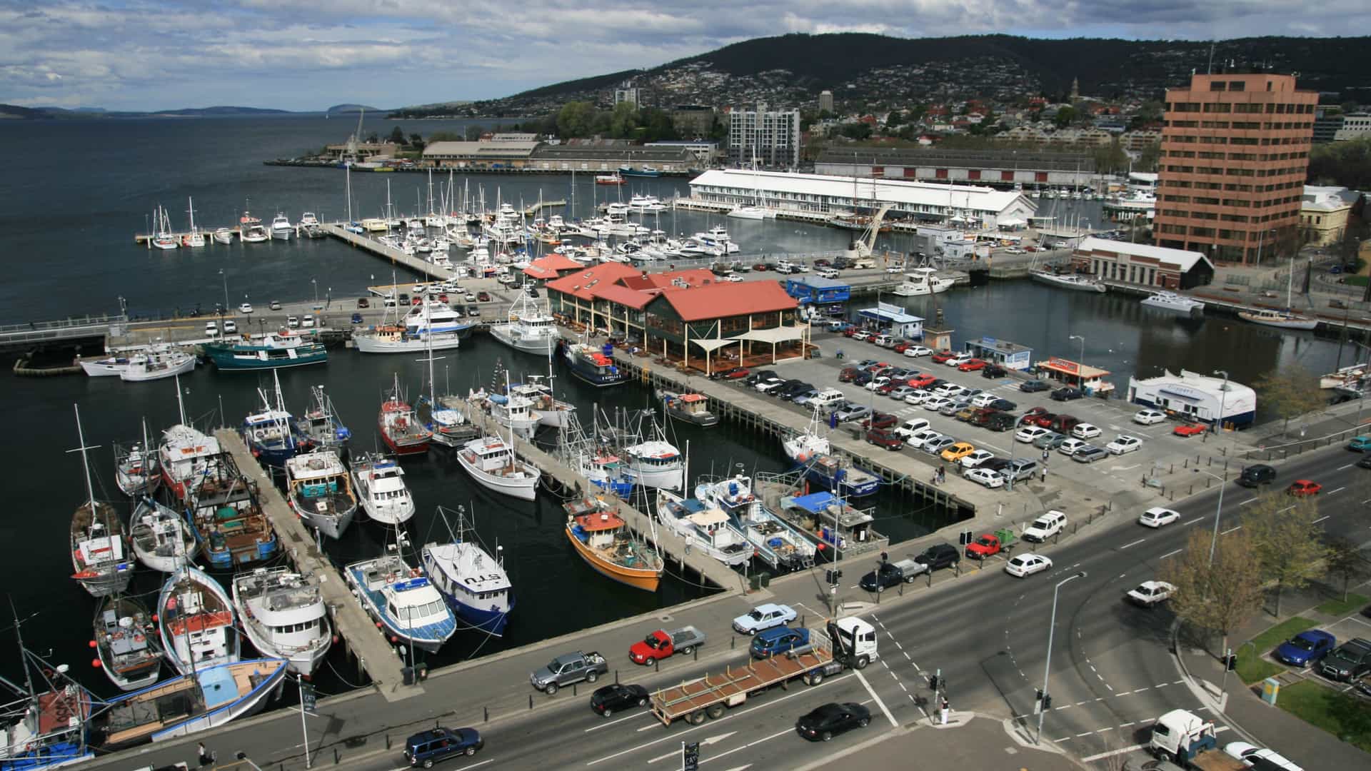 A panoramic or elevated view of the Hobart waterfront in Hobart, Tasmania, Australia, centered on Constitution Dock and Sullivans Cove. The image features several fishing boats and yachts moored in the calm water, with the historical buildings of the city's waterfront, and likely Mount Wellington (Kunanyi), visible in the background.