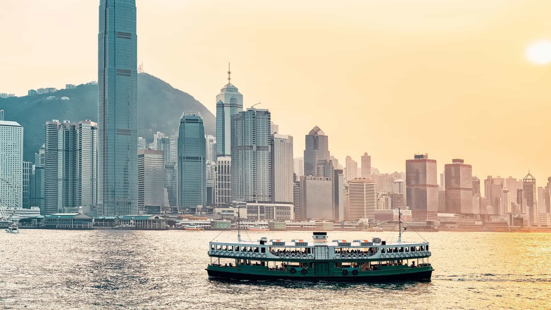 A photograph of a Star Ferry boat crossing Victoria Harbour in Hong Kong. The image shows the distinct green and white double decker ferry in the foreground. In the background, the iconic, towering skyline of Hong Kong Island is visible, with the reflection of the city lights on the water.