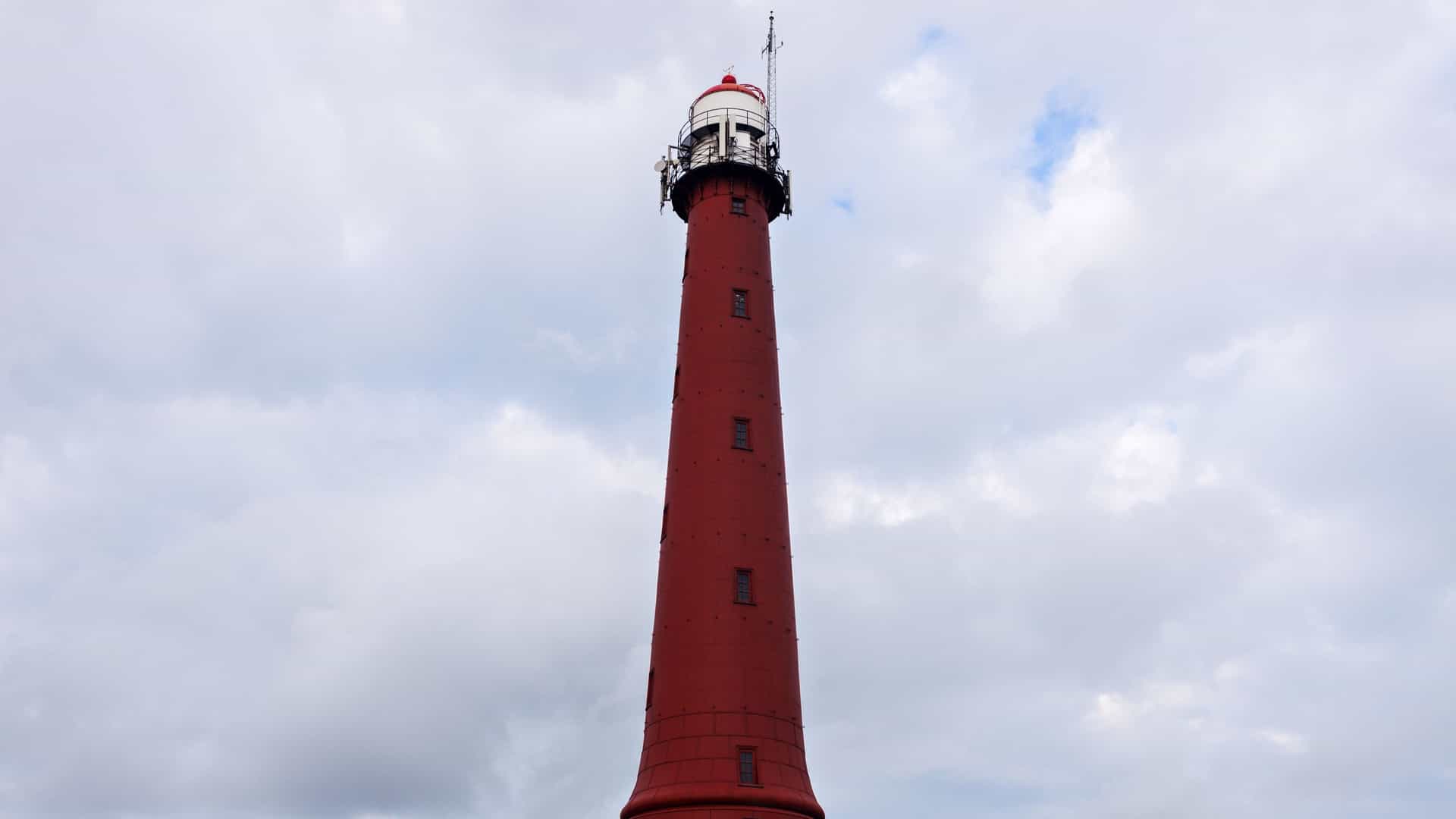 An exterior view of the Hoge vuurtoren van IJmuiden, or "high lighthouse of IJmuiden," located in the Netherlands. The lighthouse is a tall, round, reddish brown cast iron tower with a white lantern room at the top. It was built in 1878 and stands on a hill near the entrance of the North Sea Canal. The image shows the lighthouse under a blue sky, and its slim structure stands out against the surrounding landscape.