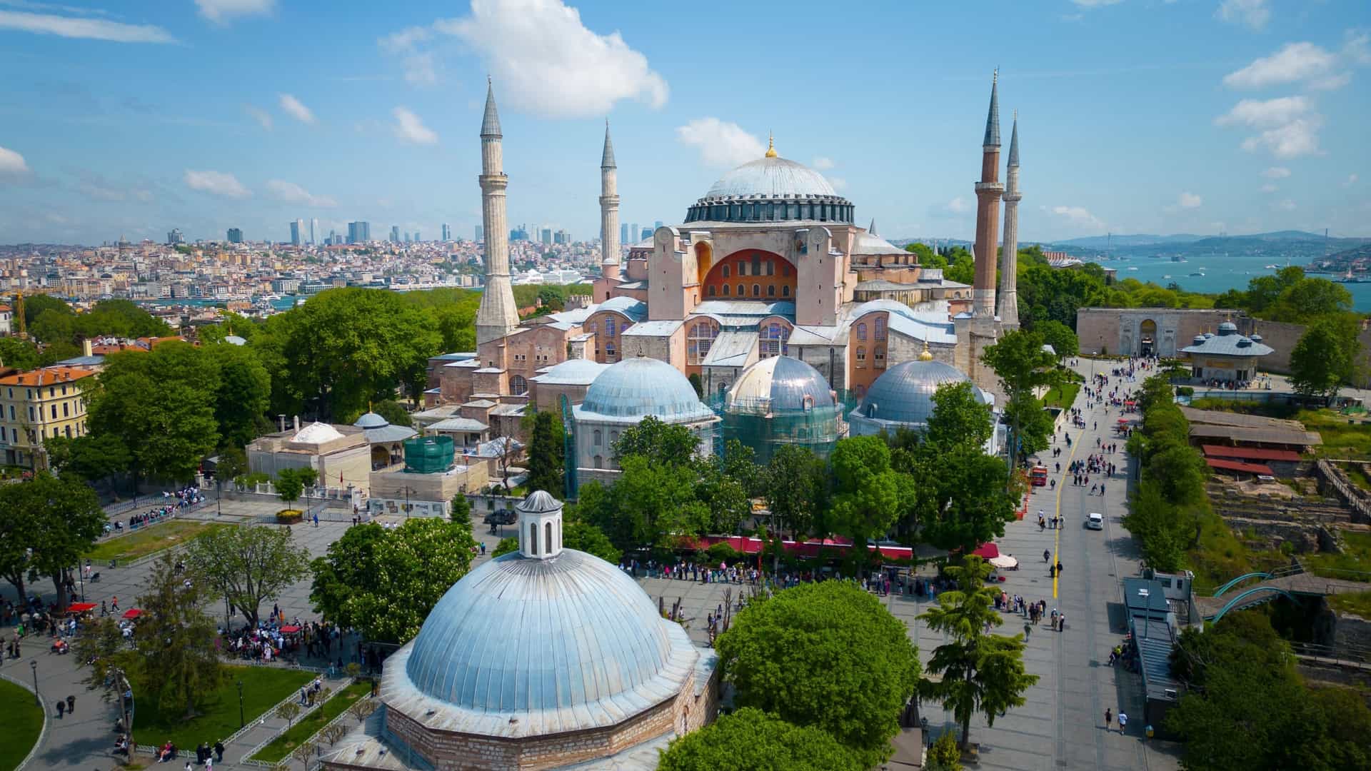 An aerial view of the Hagia Sophia Grand Mosque in Istanbul, Turkey. The Byzantine style building with its large central dome, multiple smaller domes, and minarets is surrounded by green grass and trees, and the city's urban landscape is visible in the background, all under a clear sky.