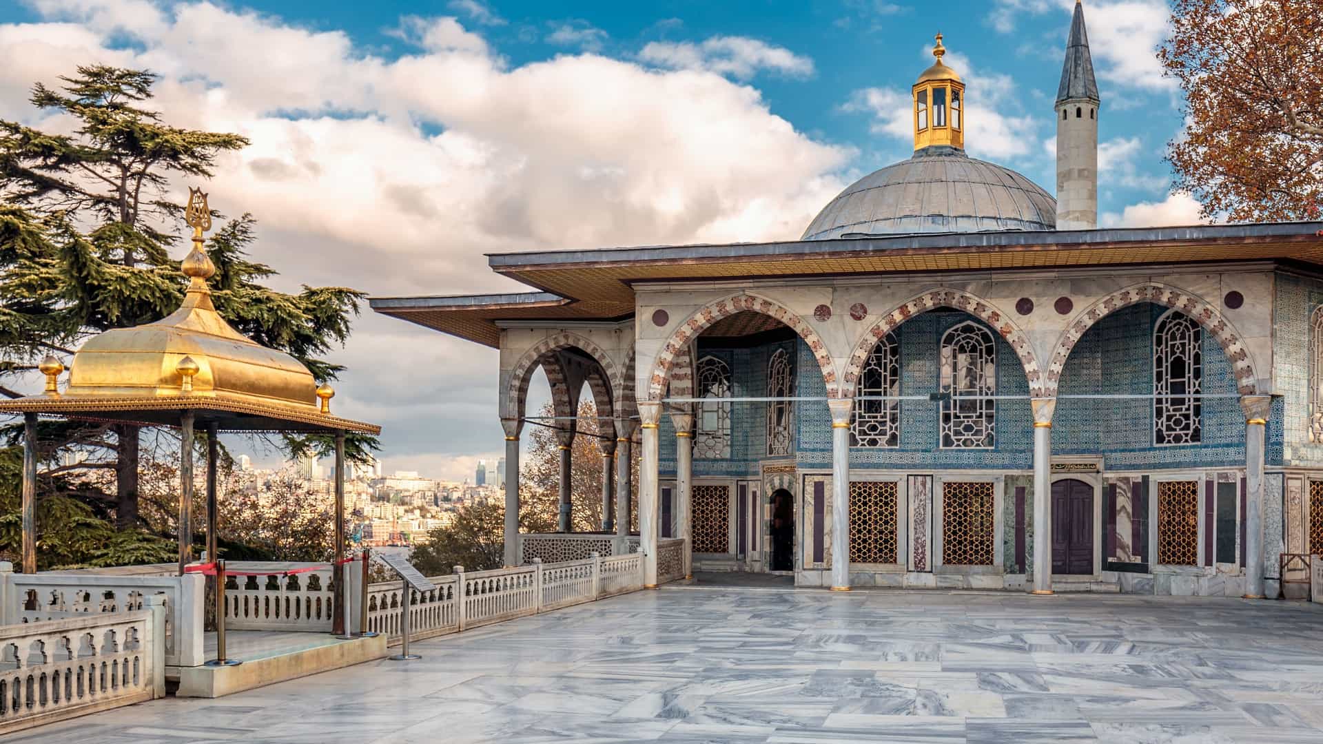 A view of the Topkapi Palace in Istanbul, Turkey, showcasing its distinctive architecture. The image captures a collection of low, red and white buildings with domed roofs, surrounded by green lawns and trees. The palace complex is a series of pavilions and courtyards, reflecting its historical role as the main residence and administrative center of the Ottoman sultans.