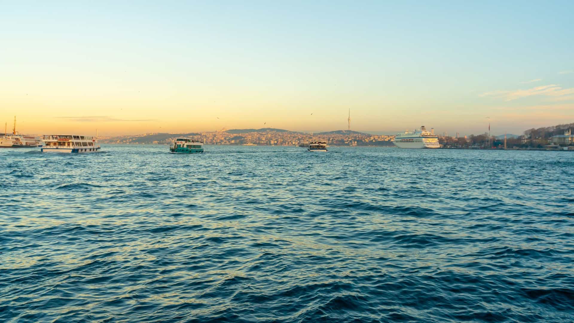 An eye level shot of a cruise ship and other vessels navigating the Bosphorus Strait in Istanbul. The cruise ship is prominently featured in the foreground, with other purpose ships, like ferries or cargo vessels, visible in the background. The scene captures the bustling maritime traffic on the famous waterway that separates the European and Asian continents, with city buildings and bridges visible on the shores.