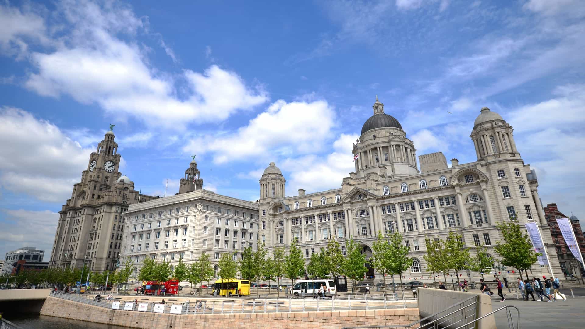A wide-angle view of the Three Graces landmark buildings on the Liverpool Pier Head, featuring the Royal Liver Building with its clock tower on the left, the Cunard Building in the center, and the Port of Liverpool Building on the right, all under a dramatic blue sky with white clouds. A street in the foreground shows red double-decker buses, other vehicles, and pedestrians, with water visible in the bottom left.