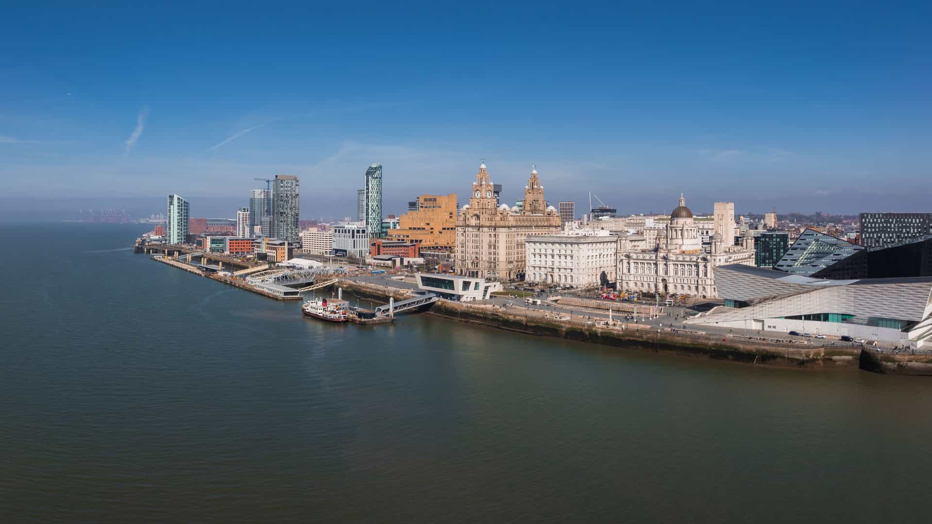 An aerial view of Liverpool's waterfront, featuring the Royal Liver Building, a prominent sandstone structure with two clock towers, each crowned by a Liver Bird statue. The River Mersey is visible in the foreground, with other city buildings surrounding the iconic landmark.