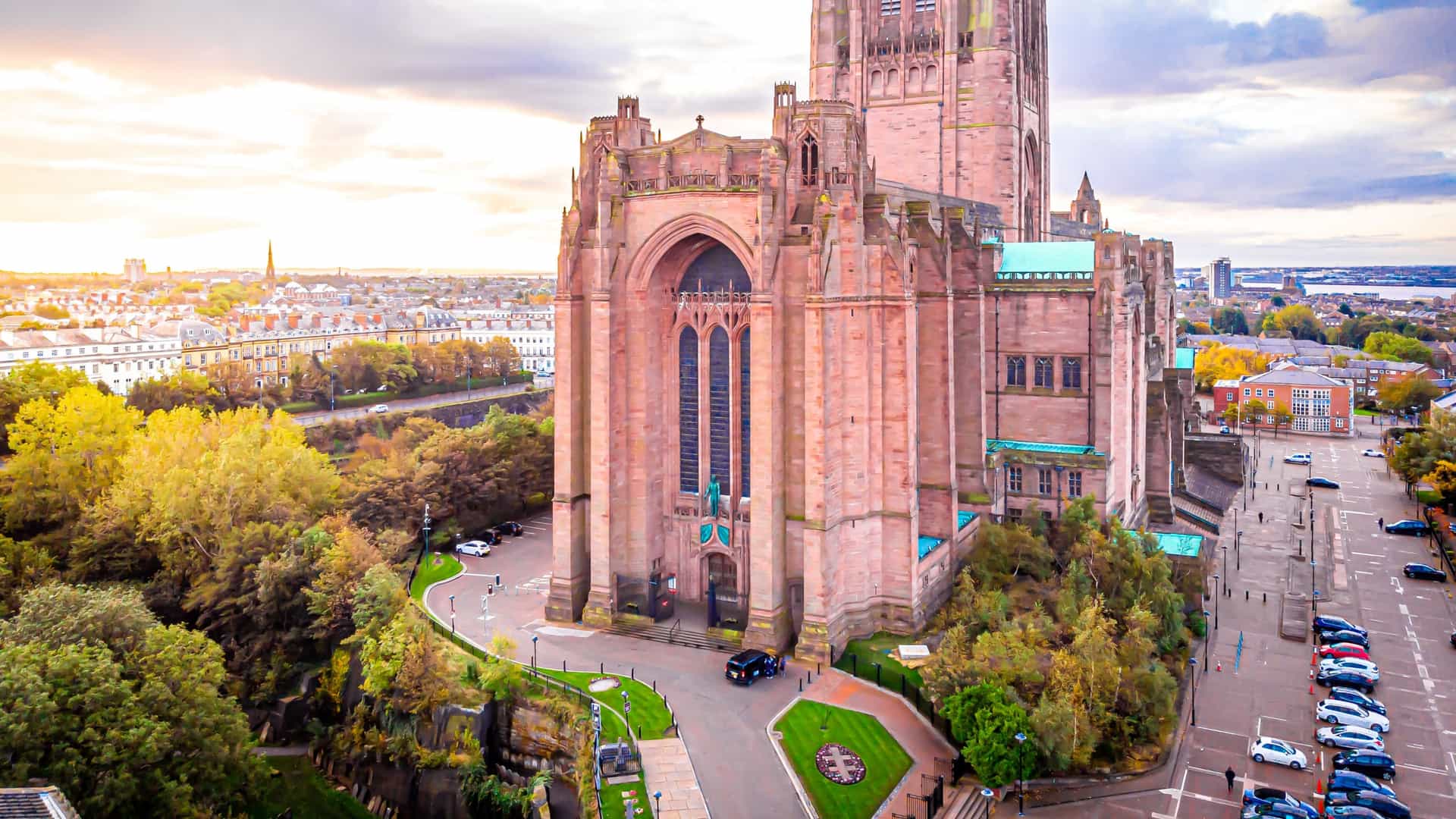 An aerial view of Liverpool Cathedral in the morning, showing the massive Gothic Revival red sandstone structure and its central tower. The cathedral is surrounded by other buildings and green spaces, with the broader city of Liverpool visible in the background under a bright morning sky.