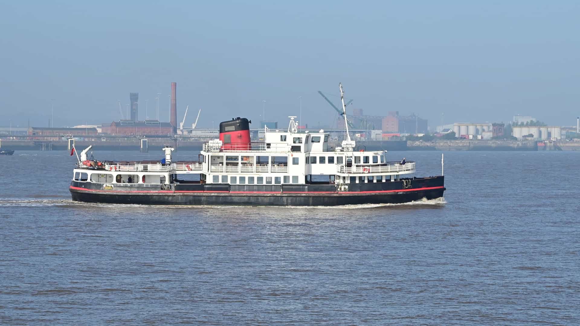 A bright, summertime view of the River Mersey with the famous Mersey Ferry sailing across the water. The iconic Liverpool skyline forms the backdrop, prominently featuring the historic 'Three Graces'—the Royal Liver Building, the Cunard Building, and the Port of Liverpool Building—under a blue sky.