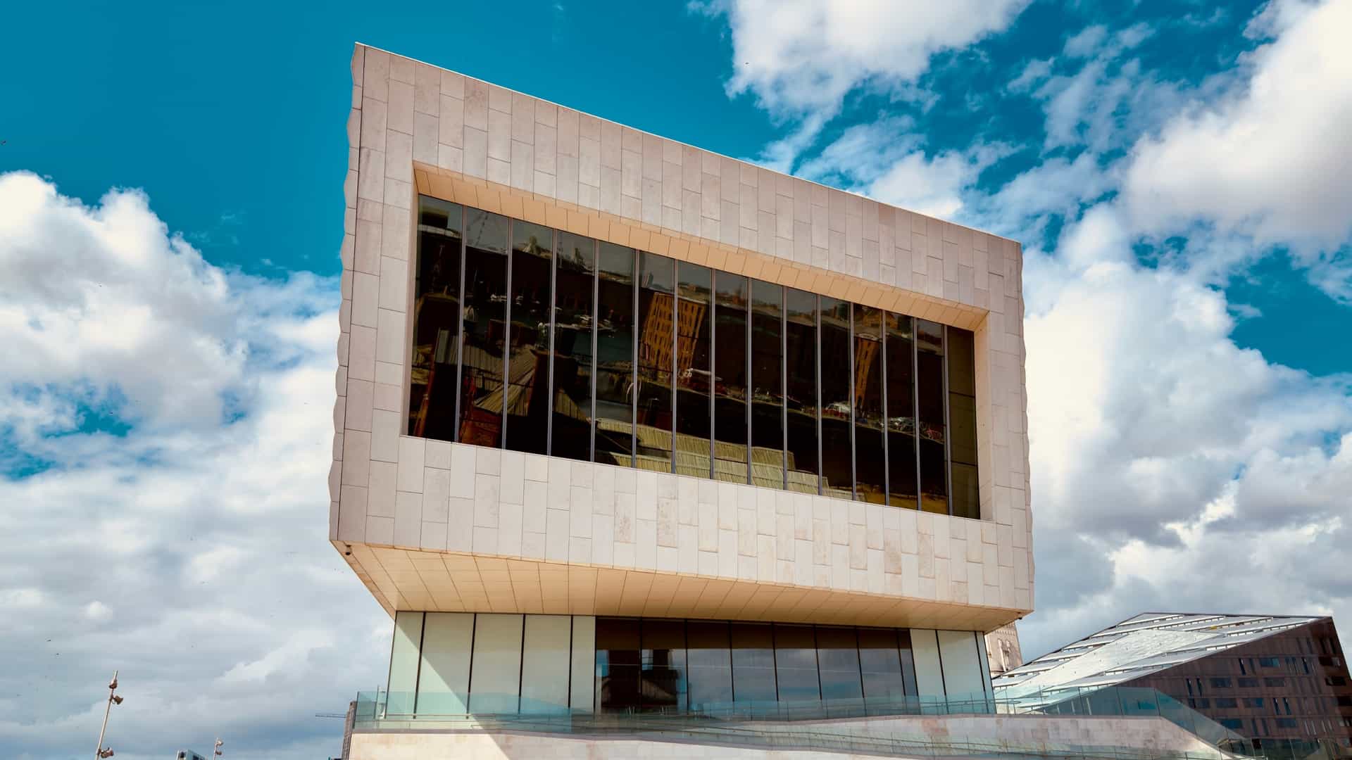 The modern, angular, and sculptural exterior of the Museum of Liverpool, with its pale stone facade and massive gabled windows, stands prominently on the Pier Head waterfront on a sunny afternoon. The building is situated next to the water, likely with a view of the famous Three Graces buildings and the River Mersey.
