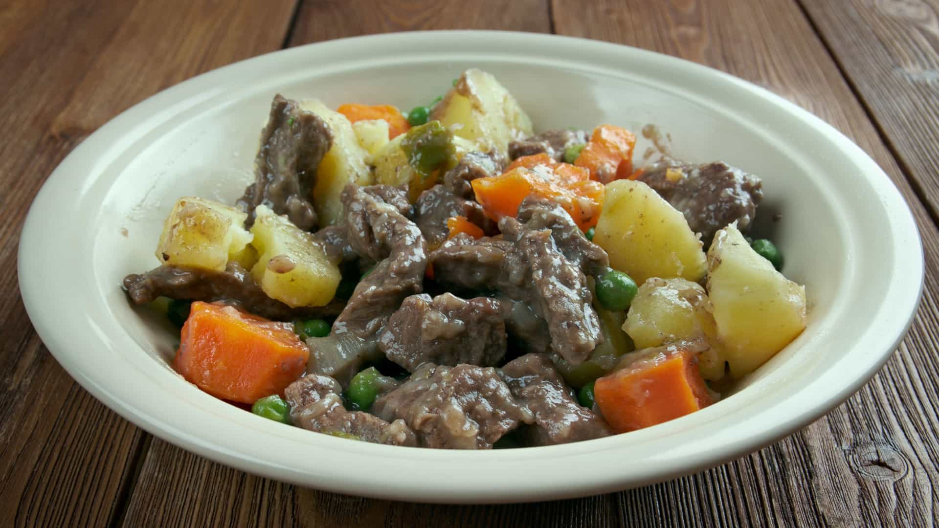 A close-up, top-down view of a serving of Scouse, a traditional Liverpudlian stew, showing a rich, thick broth filled with large, tender chunks of lamb and/or beef, potatoes, carrots, and onions, typically served with a side of pickled red cabbage or beetroot and a slice of crusty bread.