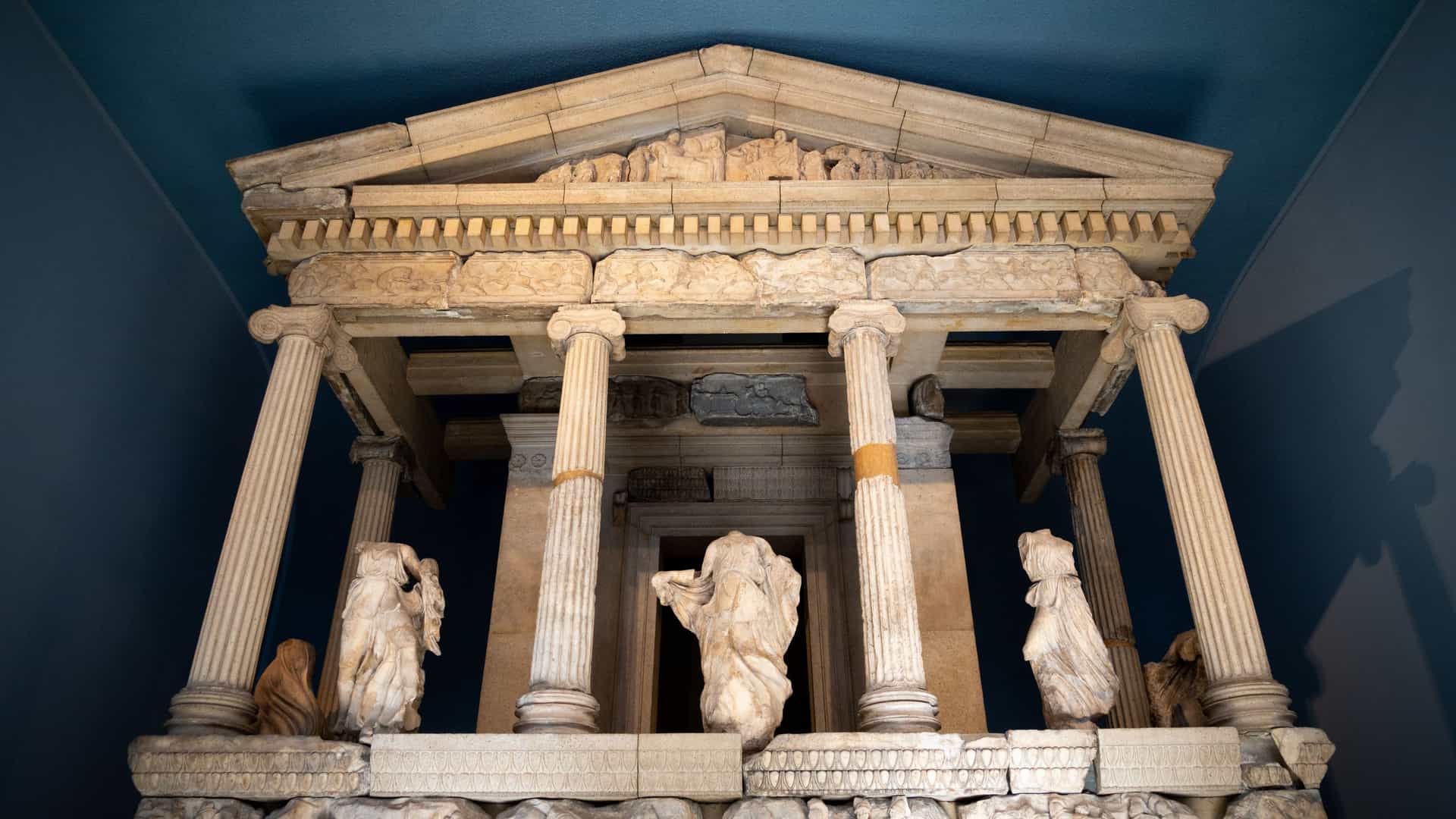 A daytime exterior view of the British Museum in London, showing its neoclassical facade with a grand entrance and symmetrical rows of tall, ornate columns under a blue sky. The museum's name is carved above the entrance.