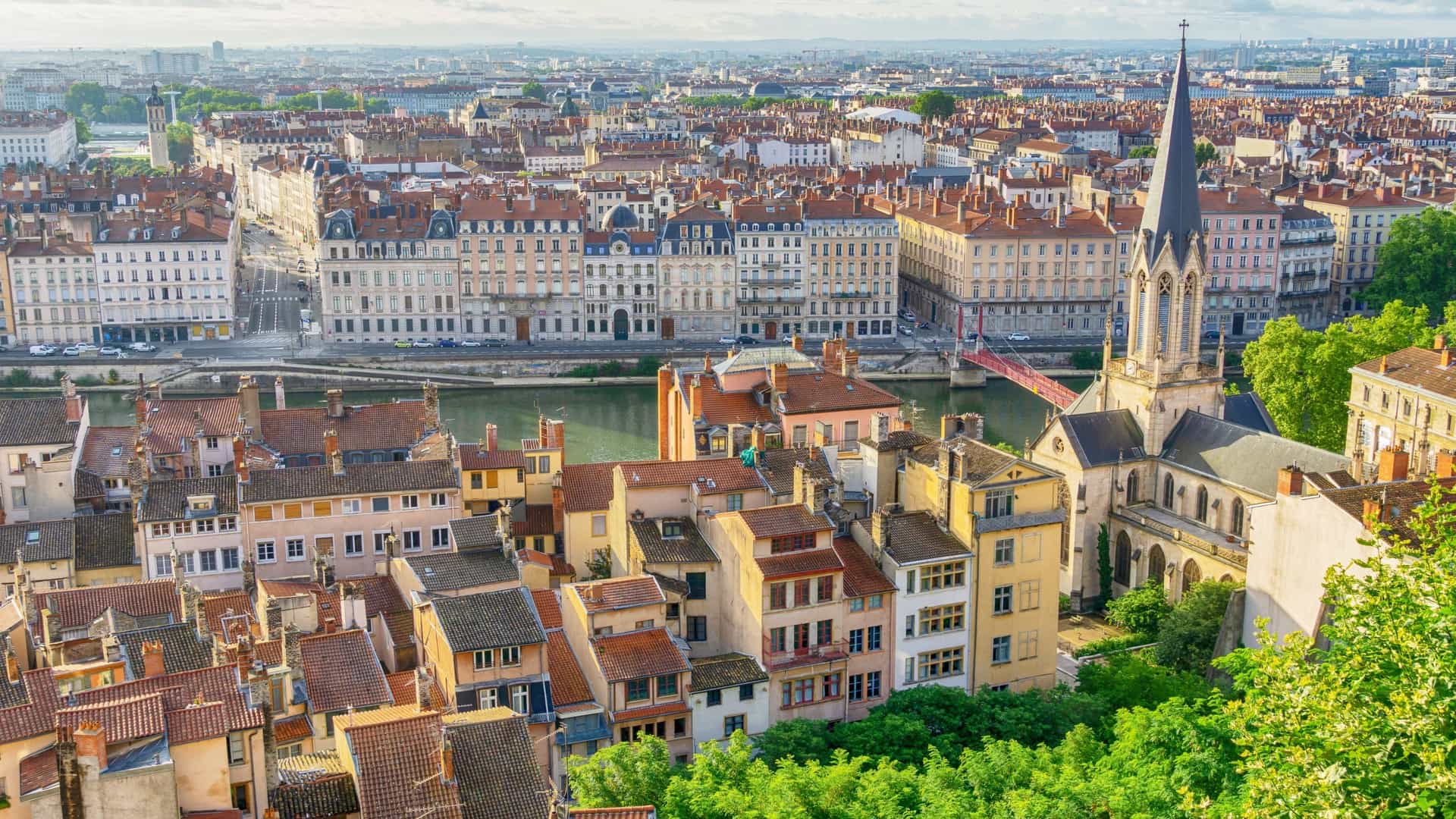 A panoramic view of Vieux Lyon, the old town of Lyon, France. The image shows a dense collection of historic, colorful buildings lining the banks of the Saône river. The Saint Jean Cathedral is visible among the buildings, and the Basilica of Notre Dame de Fourvière is prominently featured on the Fourvière Hill in the background.