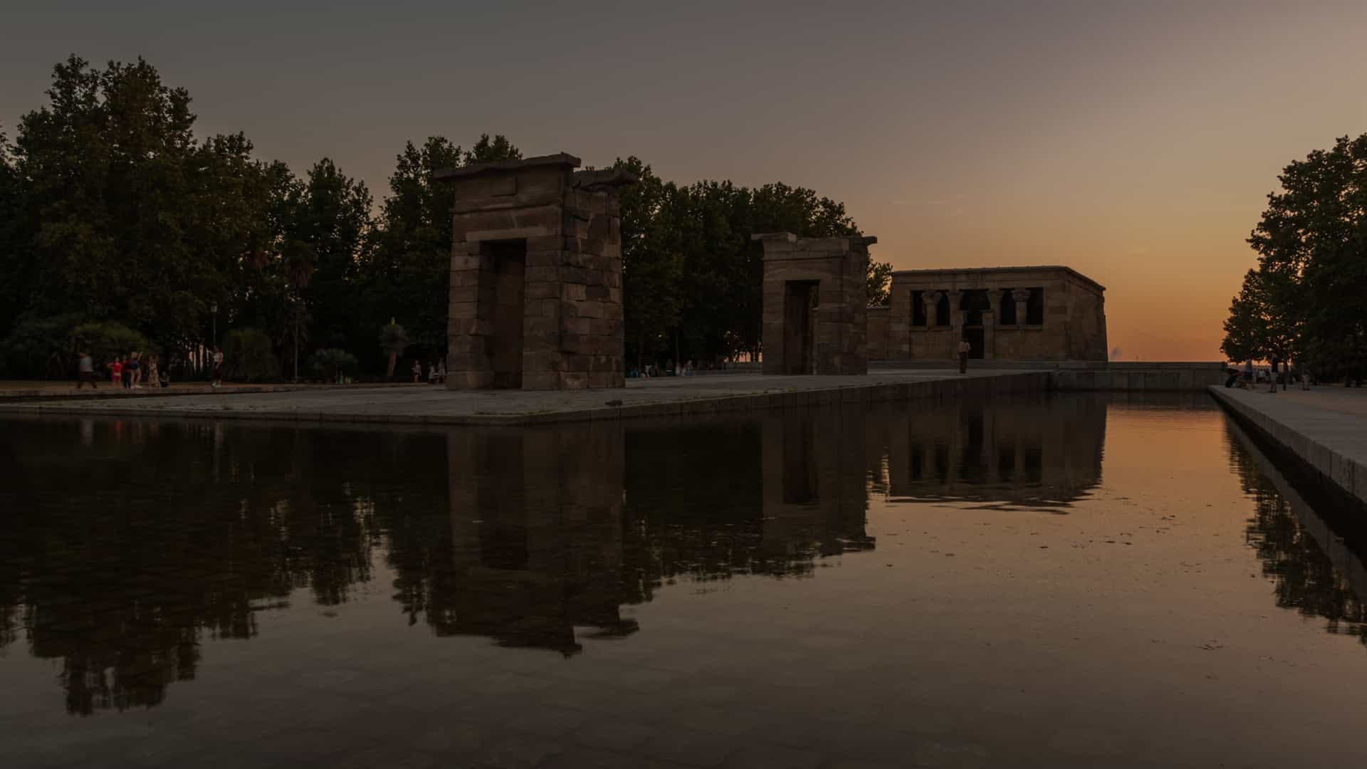 A scenic view of the Templo de Debod, an ancient Egyptian temple, located in Madrid, Spain, at sunset. The temple is surrounded by a reflecting pool, which mirrors the monument and the vibrant, colorful sky. The warm, golden light of the setting sun illuminates the stone structure, creating a striking contrast with the darkening landscape.
