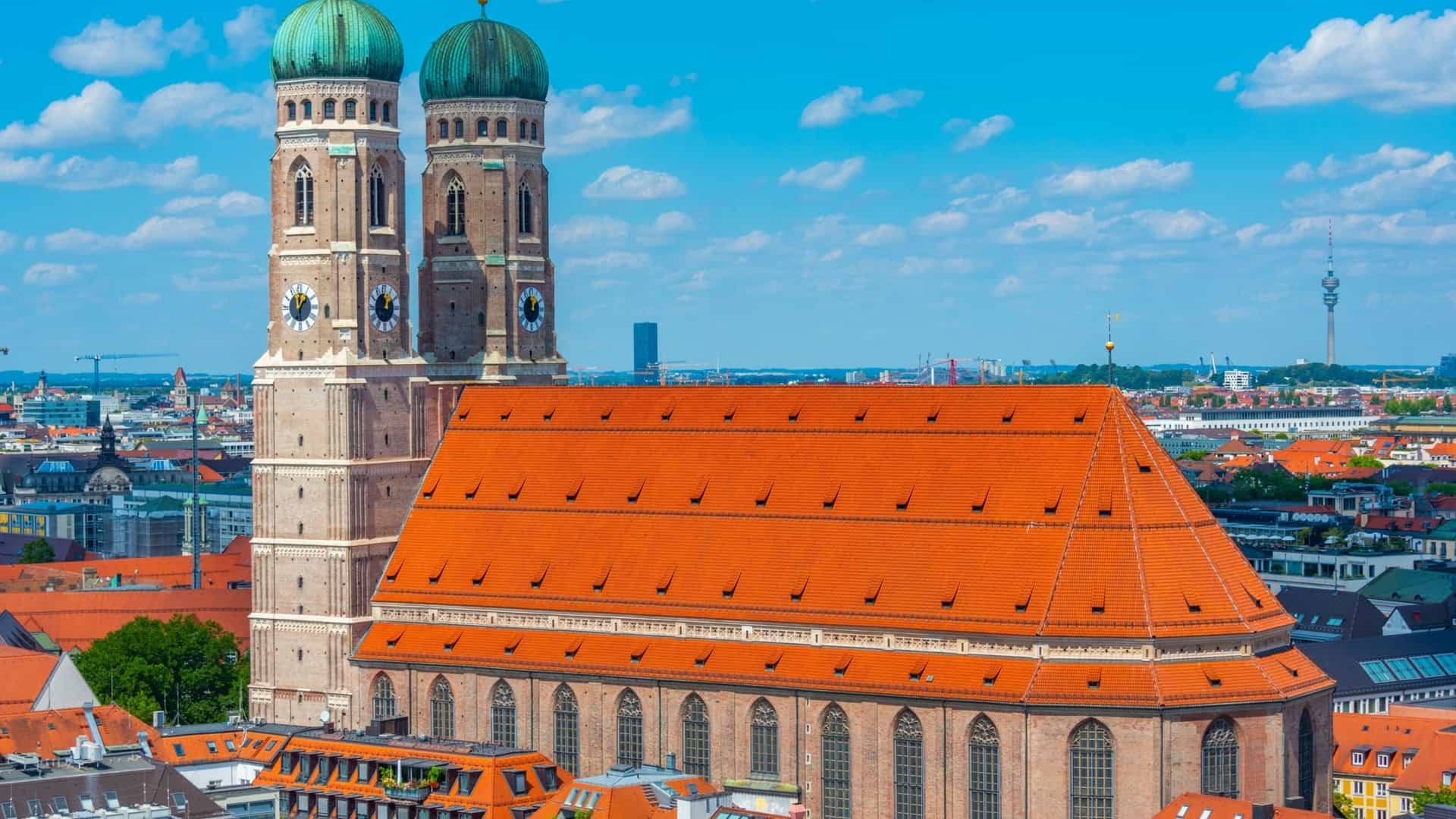 A panoramic view of Munich, Germany, with the Frauenkirche (Cathedral of Our Lady) at the center. The image shows the church's two prominent, brick towers with their distinctive green domes, surrounded by the rooftops of the city. In the background, there is a clear view of the surrounding landscape and potentially the distant Alps.