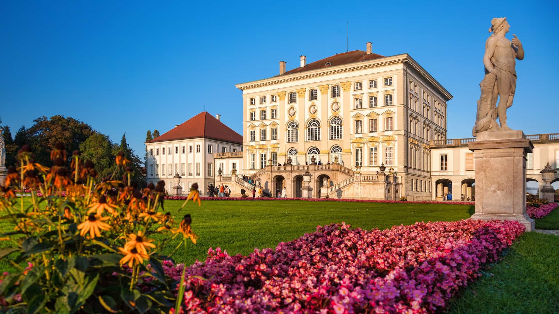 A scenic view of Nymphenburg Palace, a Baroque royal palace in Munich, Germany. The central section of the palace, a pale colored building with large windows and classical architectural details, is reflected in the still water of a canal or pond in the foreground. The image is a symmetrical composition, with the grand building and its reflection serving as the main focal point.