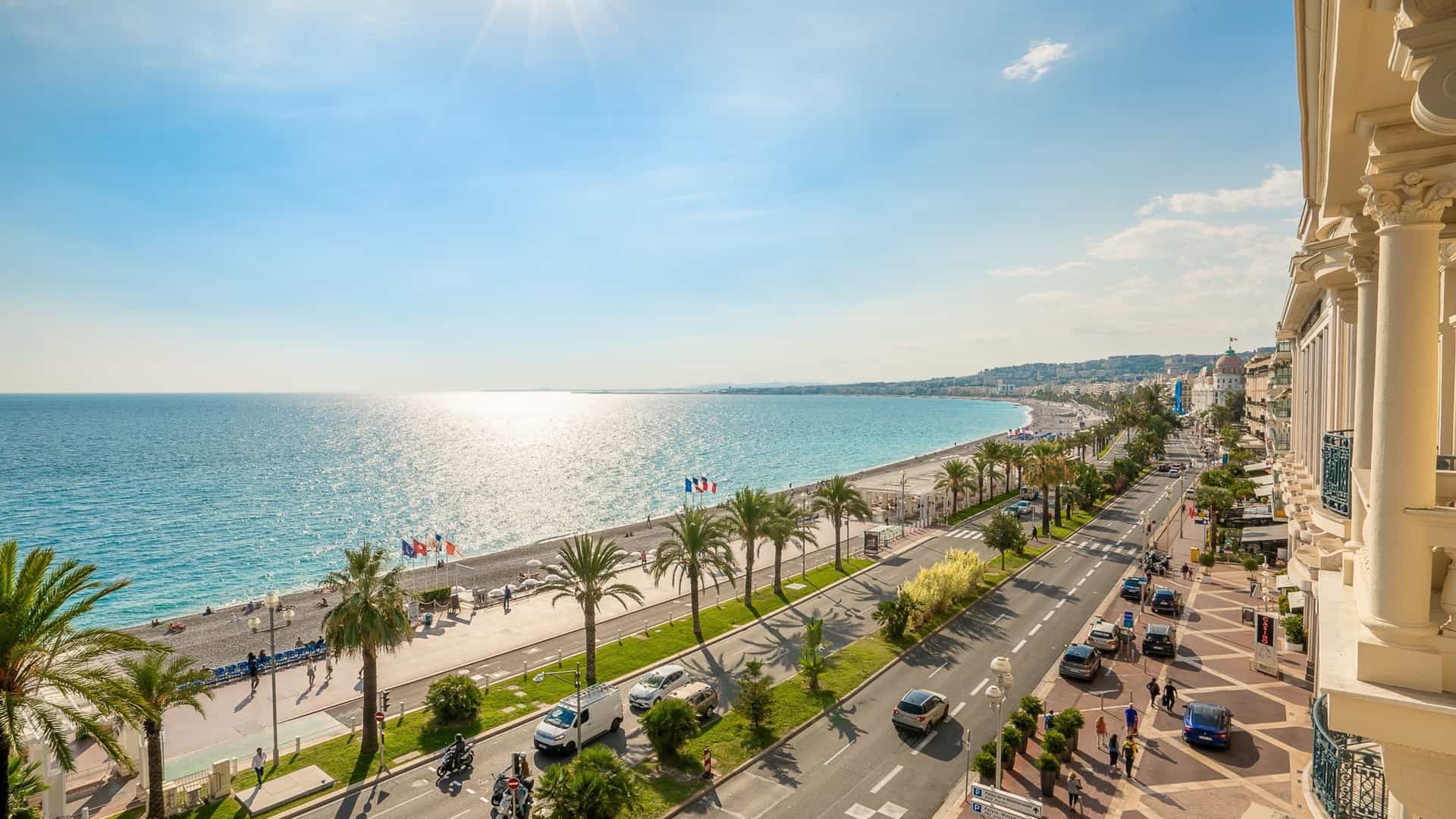 An elevated view of the Promenade des Anglais, a famous seaside promenade in Nice, France, along the Mediterranean coast. The image shows the wide, palm tree lined walkway, with the bright blue sea on one side and a row of elegant buildings on the other.